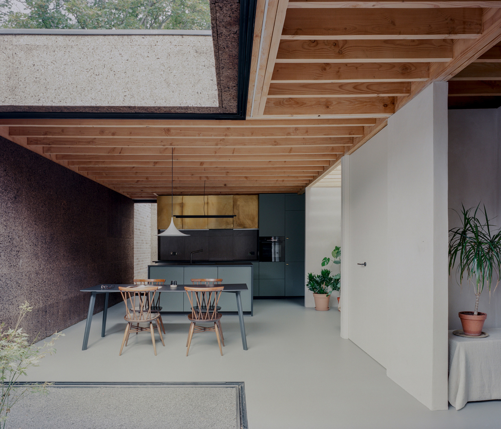Wide view of the kitchen and dining space with timber joist ceiling, cork wall, green cabinets and a rooflight over a planted void