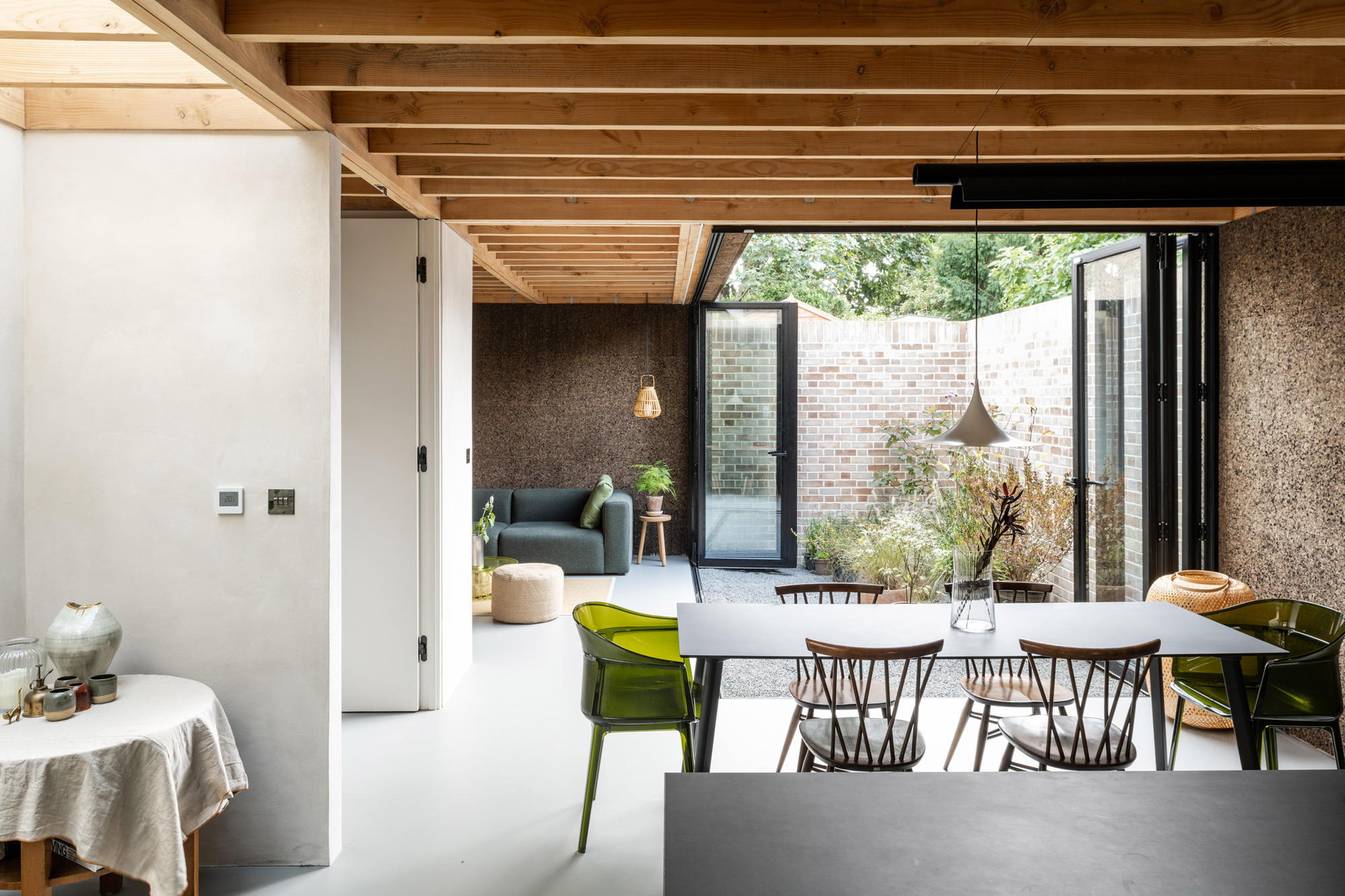 Open-plan dining area with exposed timber joists, folding glass doors and a brick-walled courtyard planted with grasses in daylight