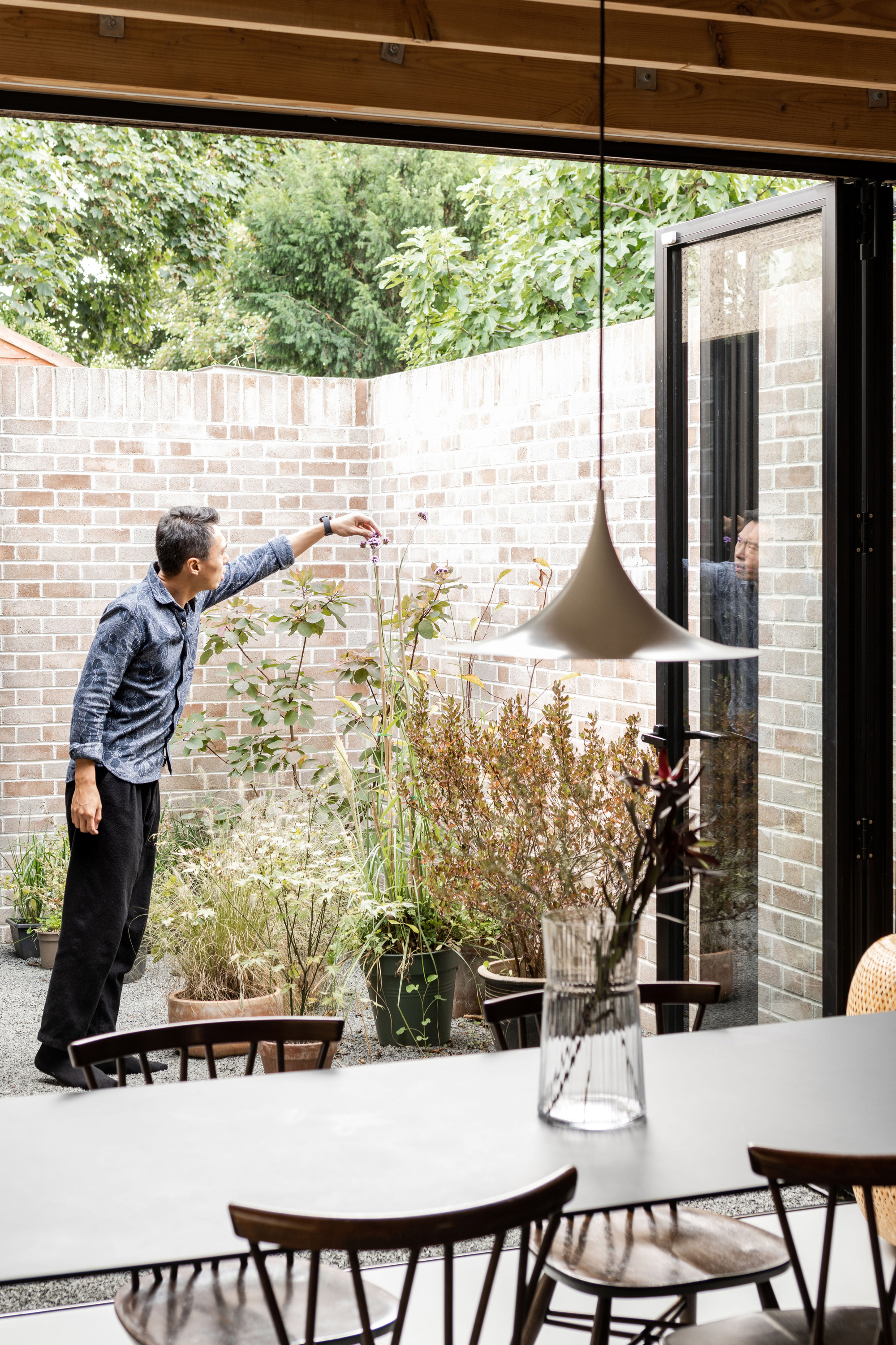 Man reaching toward a potted plant in a brick courtyard, seen past a dining table and pendant lamp