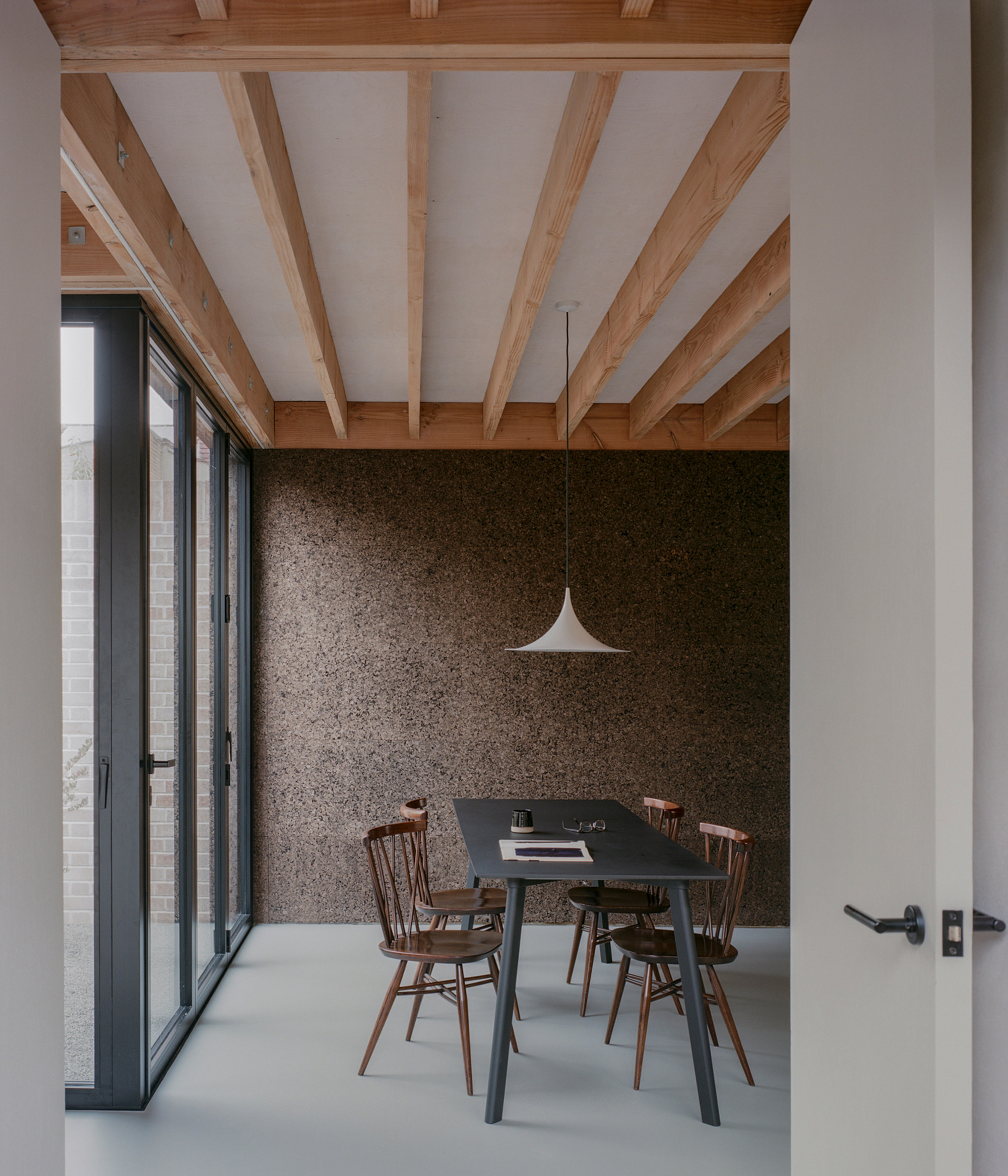 Dining table with timber chairs against a cork-clad wall, beneath exposed joists and a white pendant lamp, with glazed doors at left