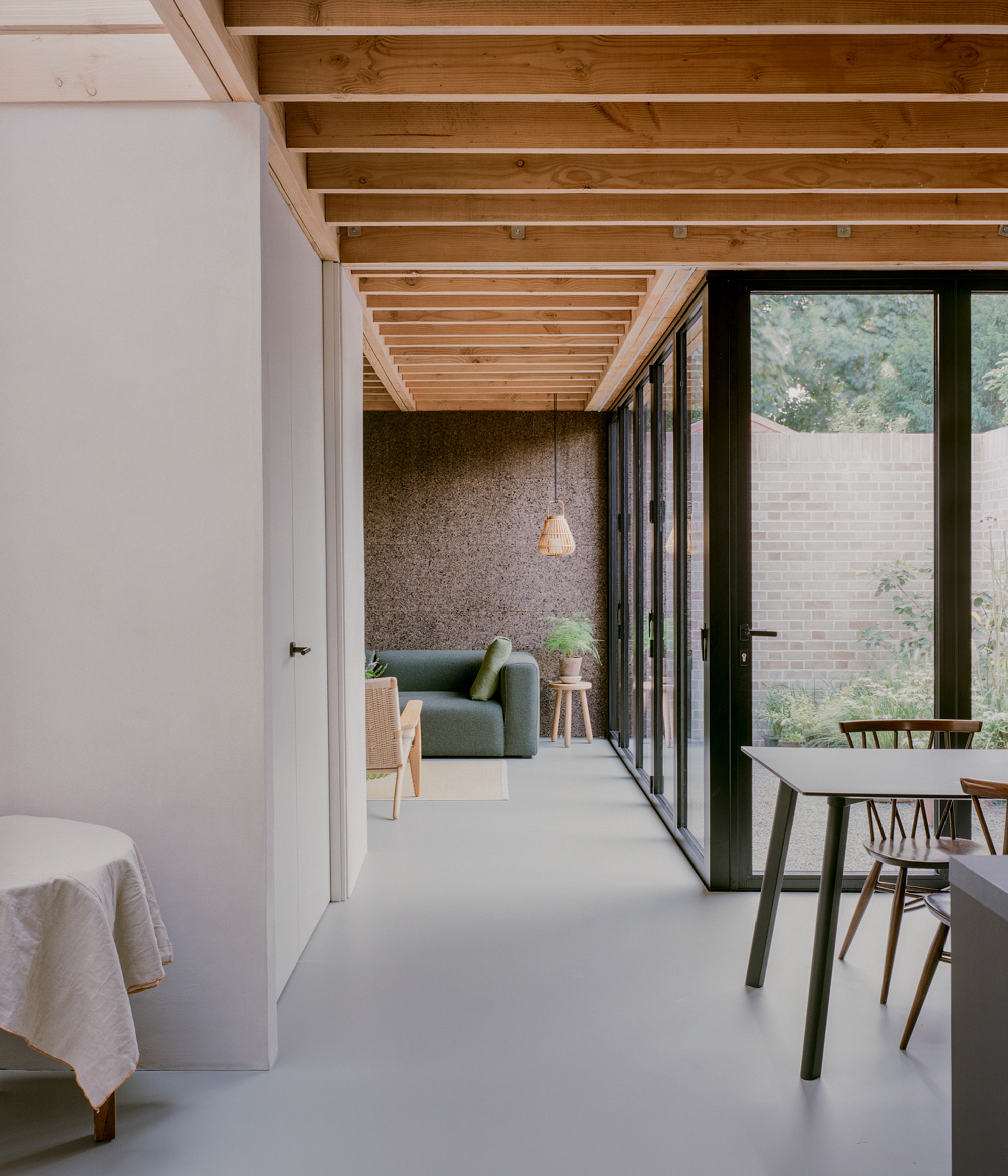 View along the glazed wall toward a green sofa and cork-clad alcove, with garden greenery reflected in the folding doors