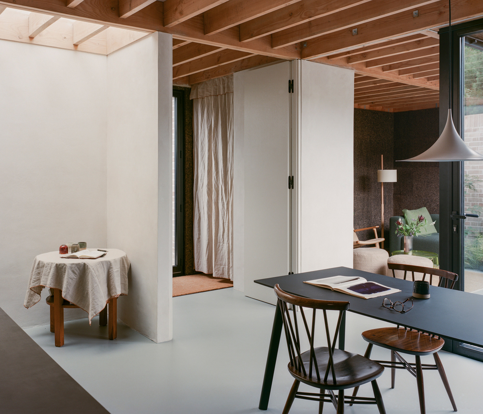 Dining table with open book and spectacles in the foreground, a linen-draped side table and plastered partition walls beyond under timber joists