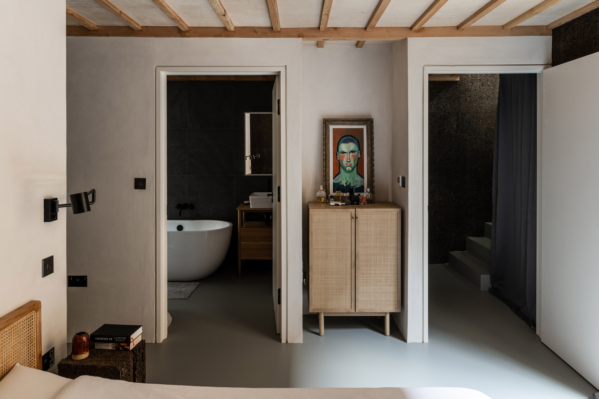 Bedroom view toward a rattan cabinet and framed portrait, with an ensuite bathtub and a cork-lined stair passage on either side