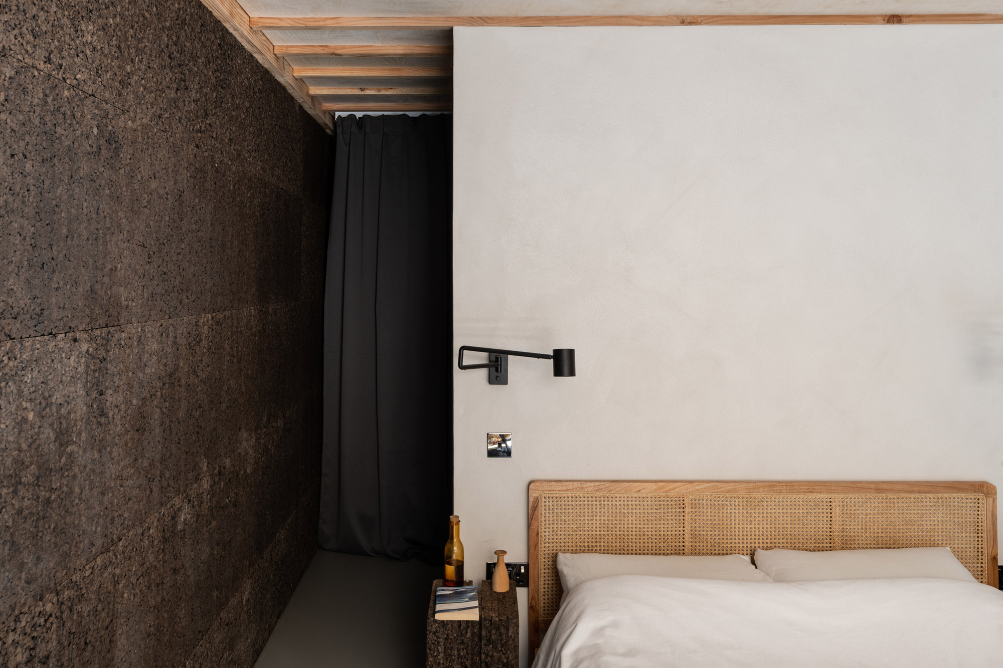 Bedside corner with a rattan headboard, black swing-arm lamp and dark cork wall beside a heavy charcoal curtain