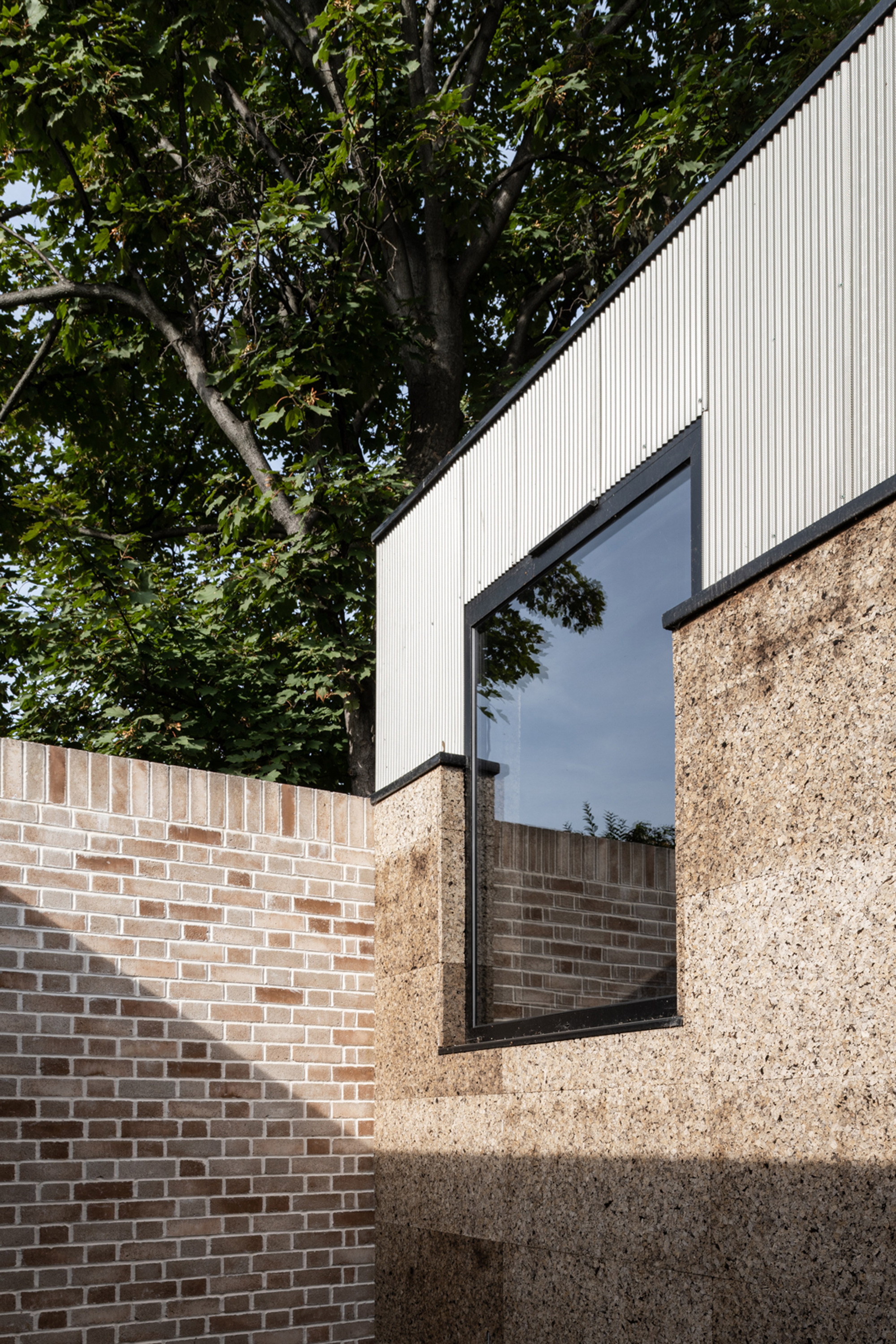 Exterior corner where corrugated metal cladding, a cork-textured wall and a brick garden wall meet beneath a leafy tree in sunlight