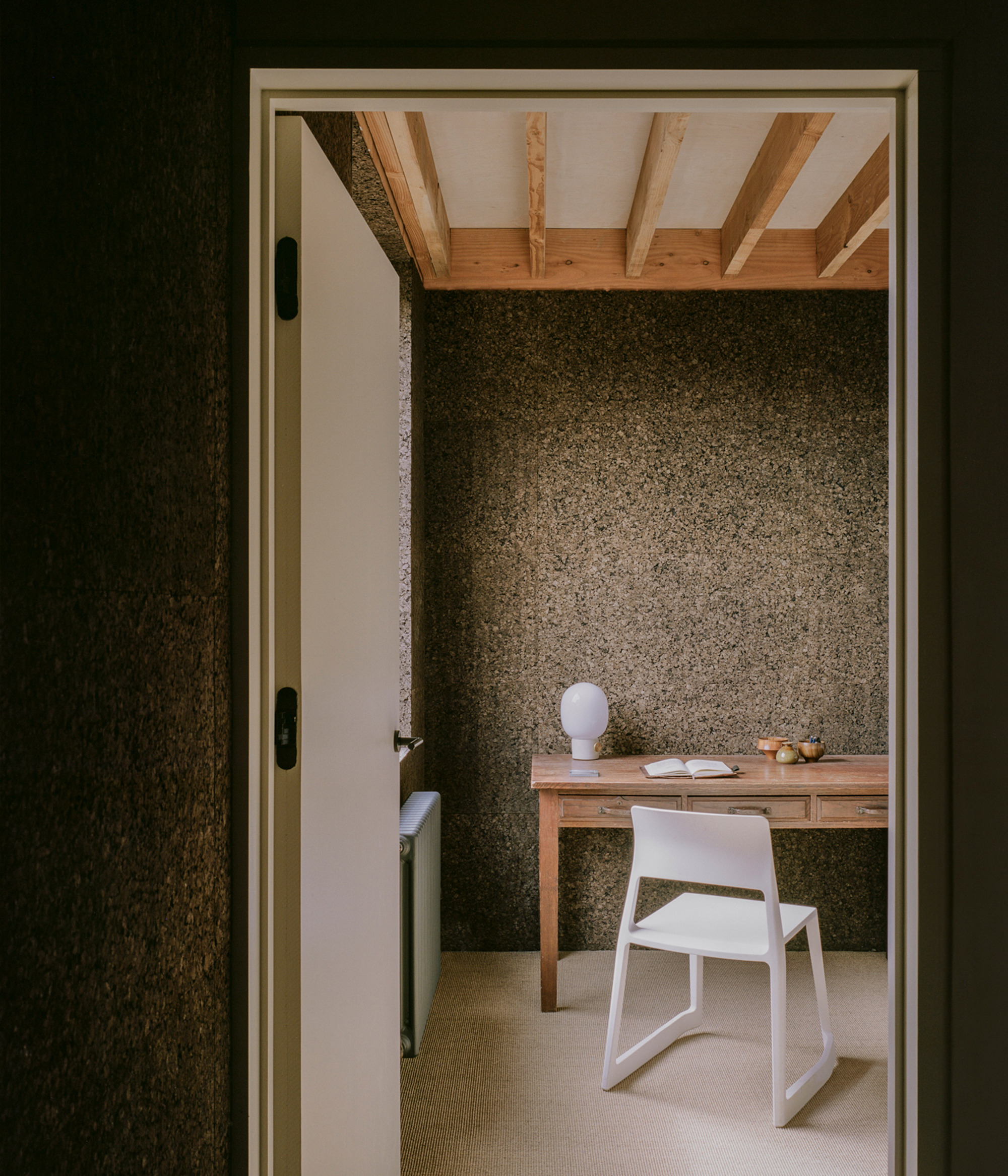 Doorway view into a small study with a timber desk, white chair and cork walls under exposed ceiling joists