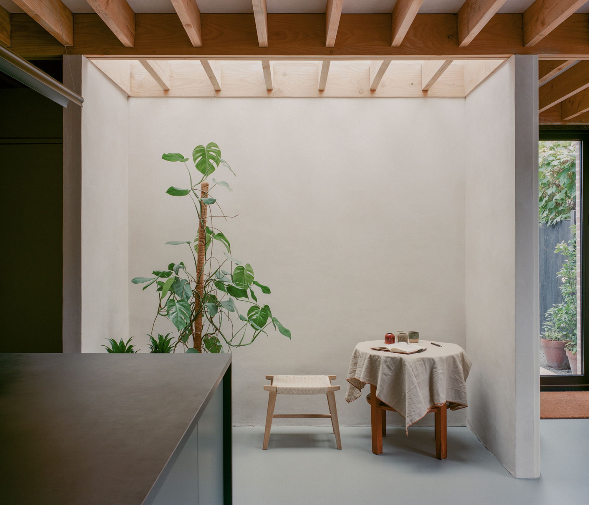 Skylit dining alcove with a linen-covered table, woven stool and tall monstera, a garden glimpsed through the glazed door