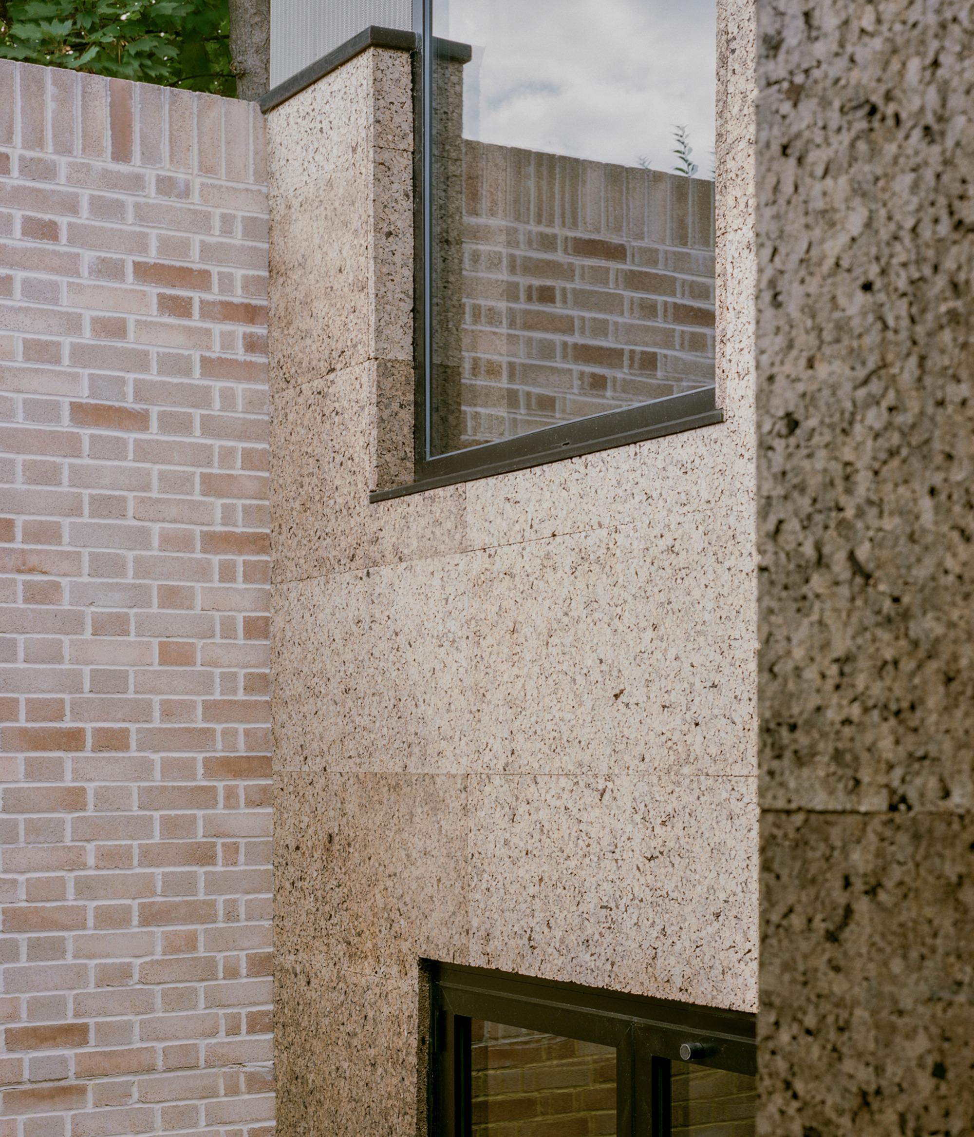 Corner of cork-clad walls beside a pale brick wall, with a steel-framed window reflecting brick under a cloudy sky