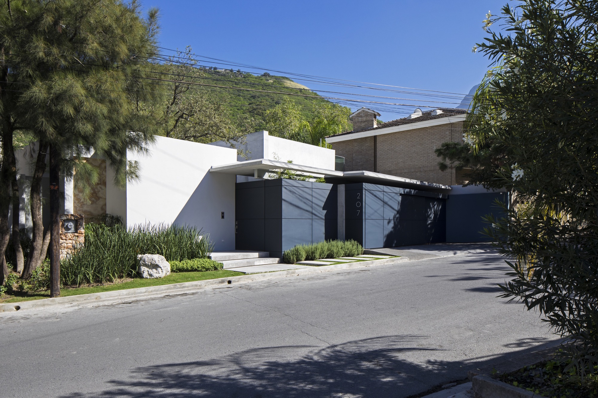 Street facade of white walls and dark grey panel gates, framed by pine and shrub foliage in bright midday sun