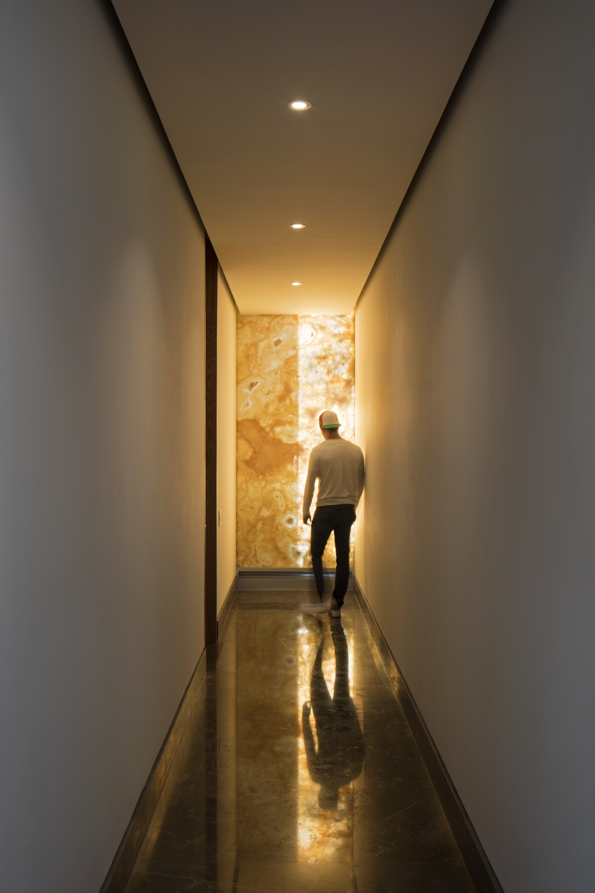 Narrow hallway ending in a backlit onyx wall, with a man walking toward it and reflected in the glossy floor