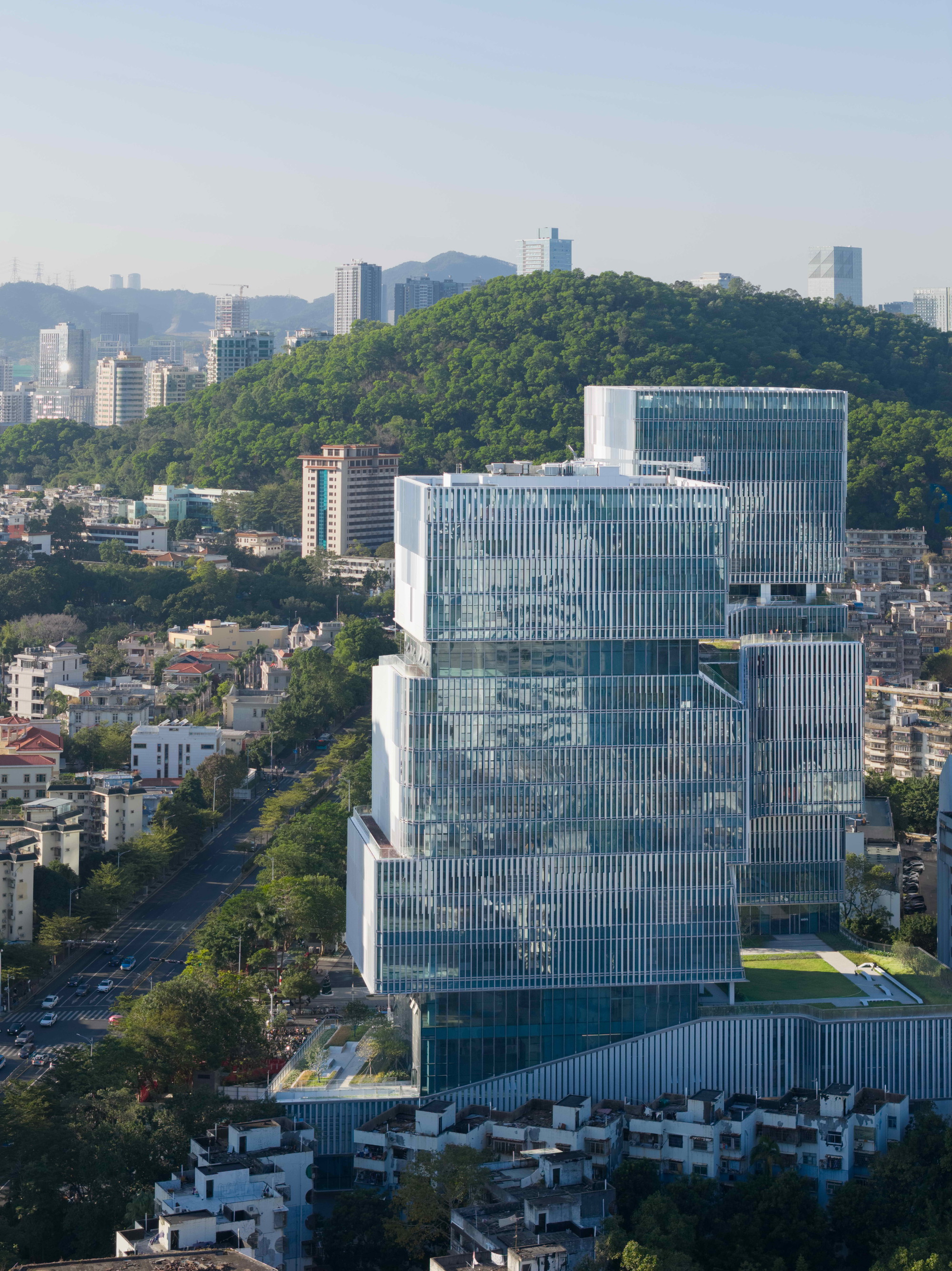 Aerial view of the offset glass towers rising above low rooftops, with a forested hill behind in clear daylight