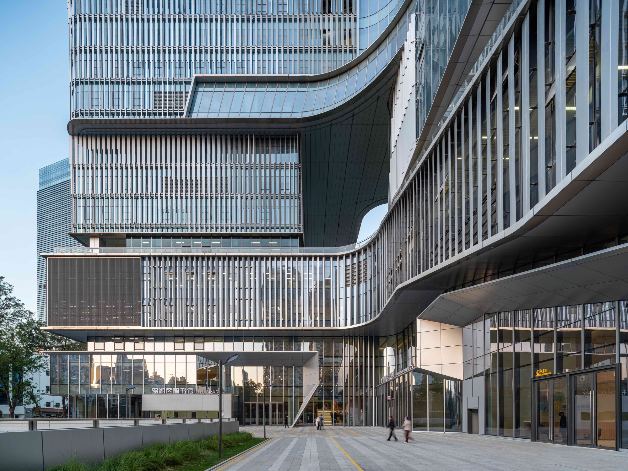 Curving underside of the cantilevered volumes above a paved plaza, with pedestrians walking past the glazed entrance