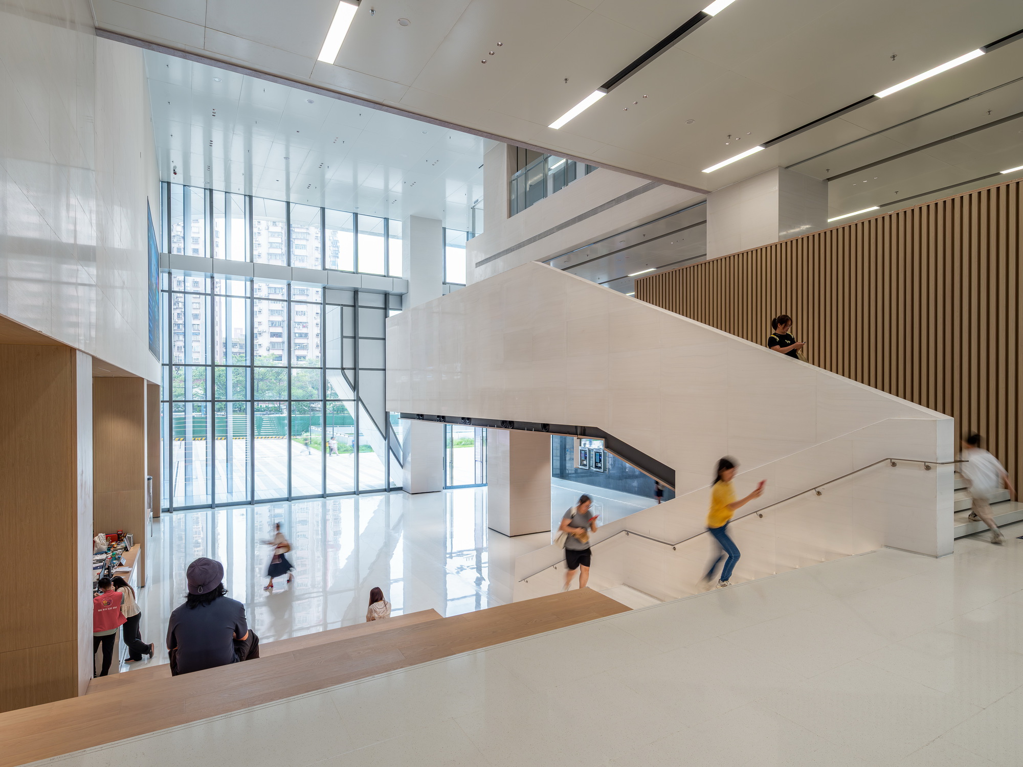 Interior stair hall with white stone flight, timber slat wall and full-height glazing, visitors moving past in daylight