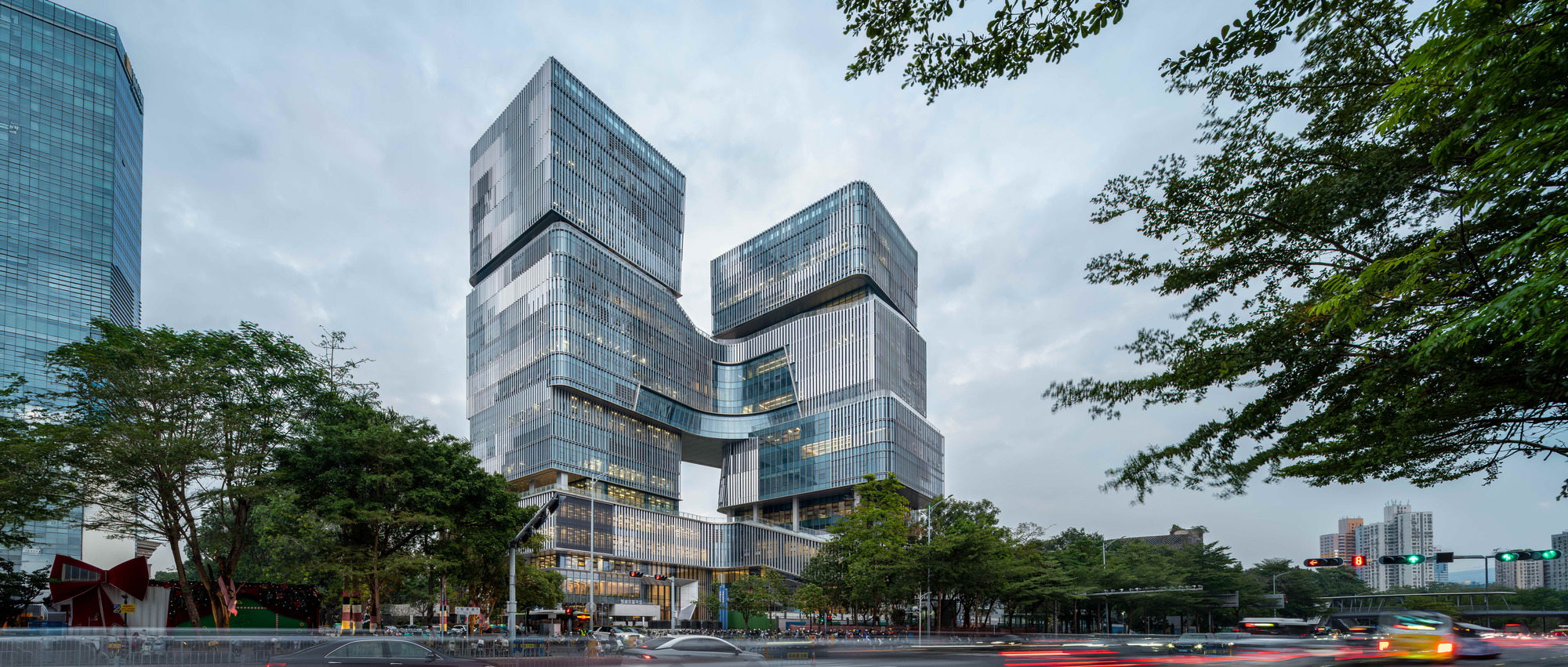 Two stacked glass towers joined by a bridge, framed by street trees under an overcast sky with blurred traffic below