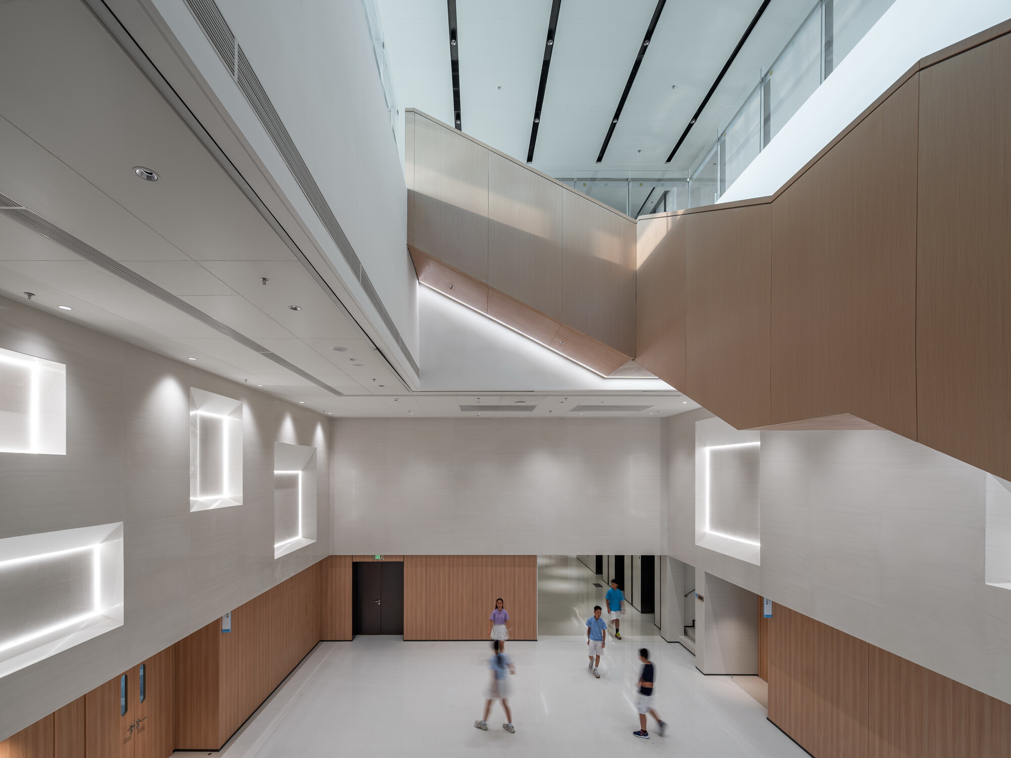 Atrium with timber-clad stair soffit and recessed light-framed wall niches, students crossing the pale floor in motion blur
