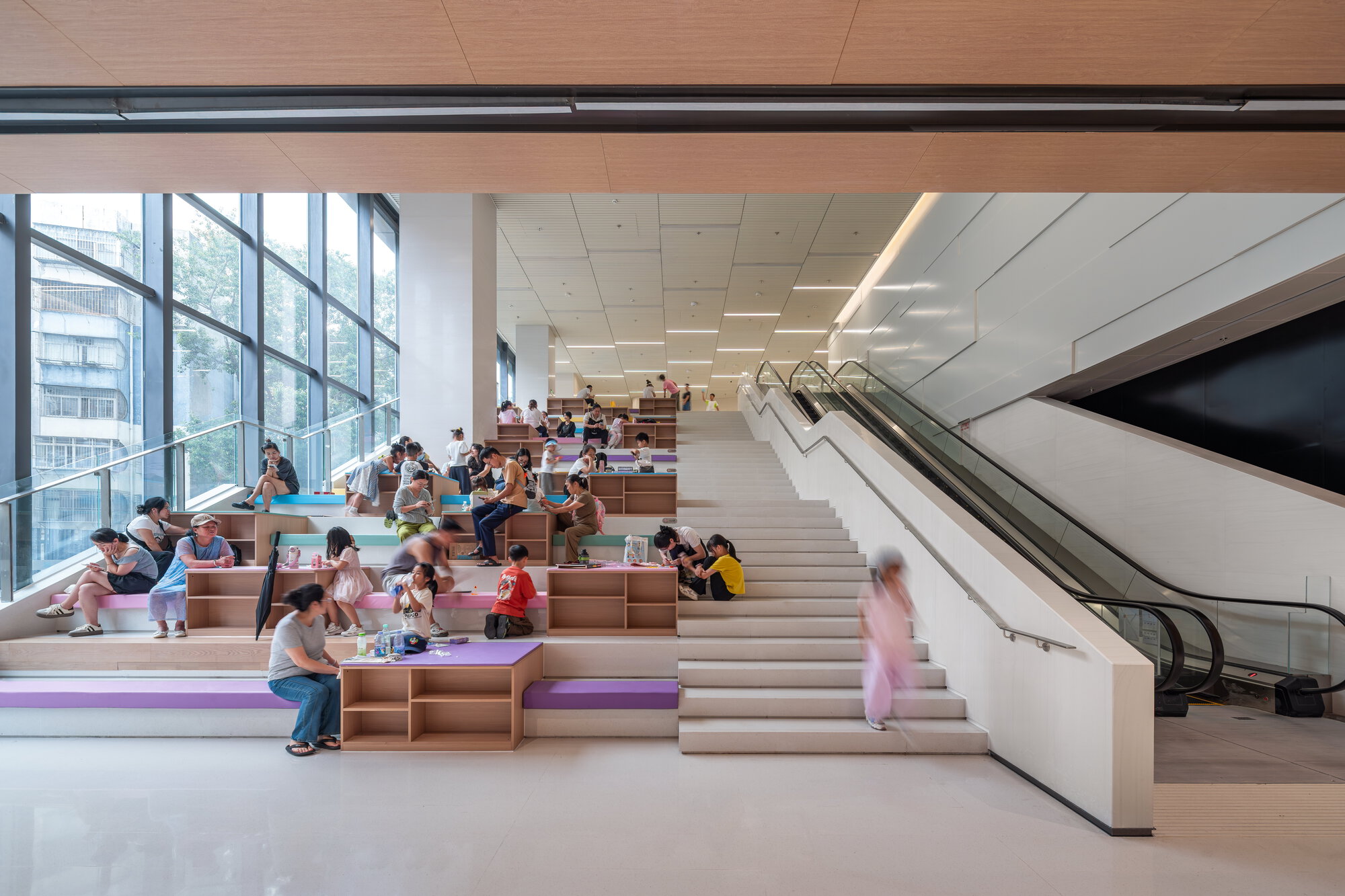 Wide stepped seating with timber shelves and purple cushions beside a stair and escalator, readers gathered along a glazed wall