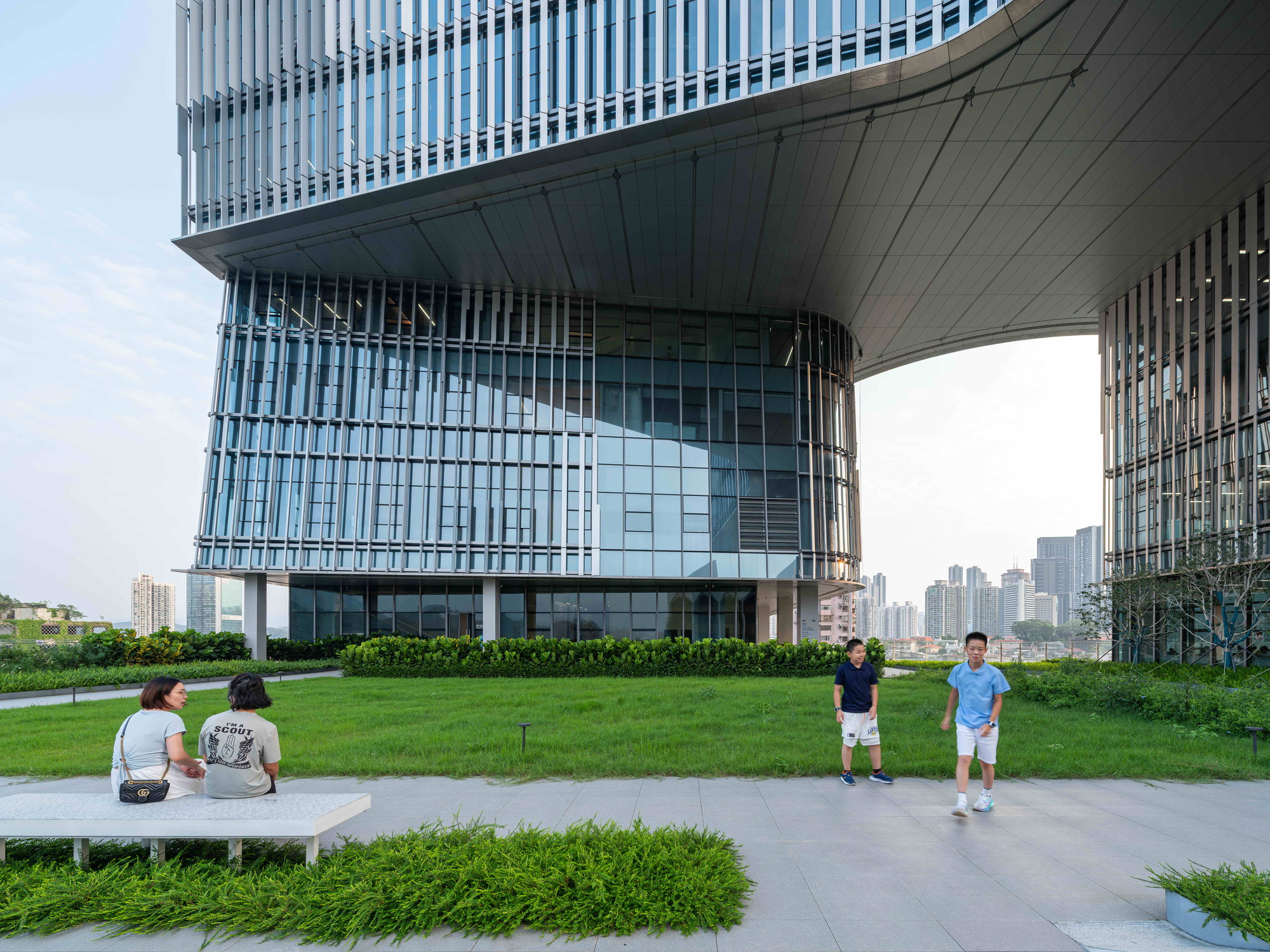 Rooftop lawn and planted beds beneath the curved underside of the tower, two boys walking past a seated pair on a bench