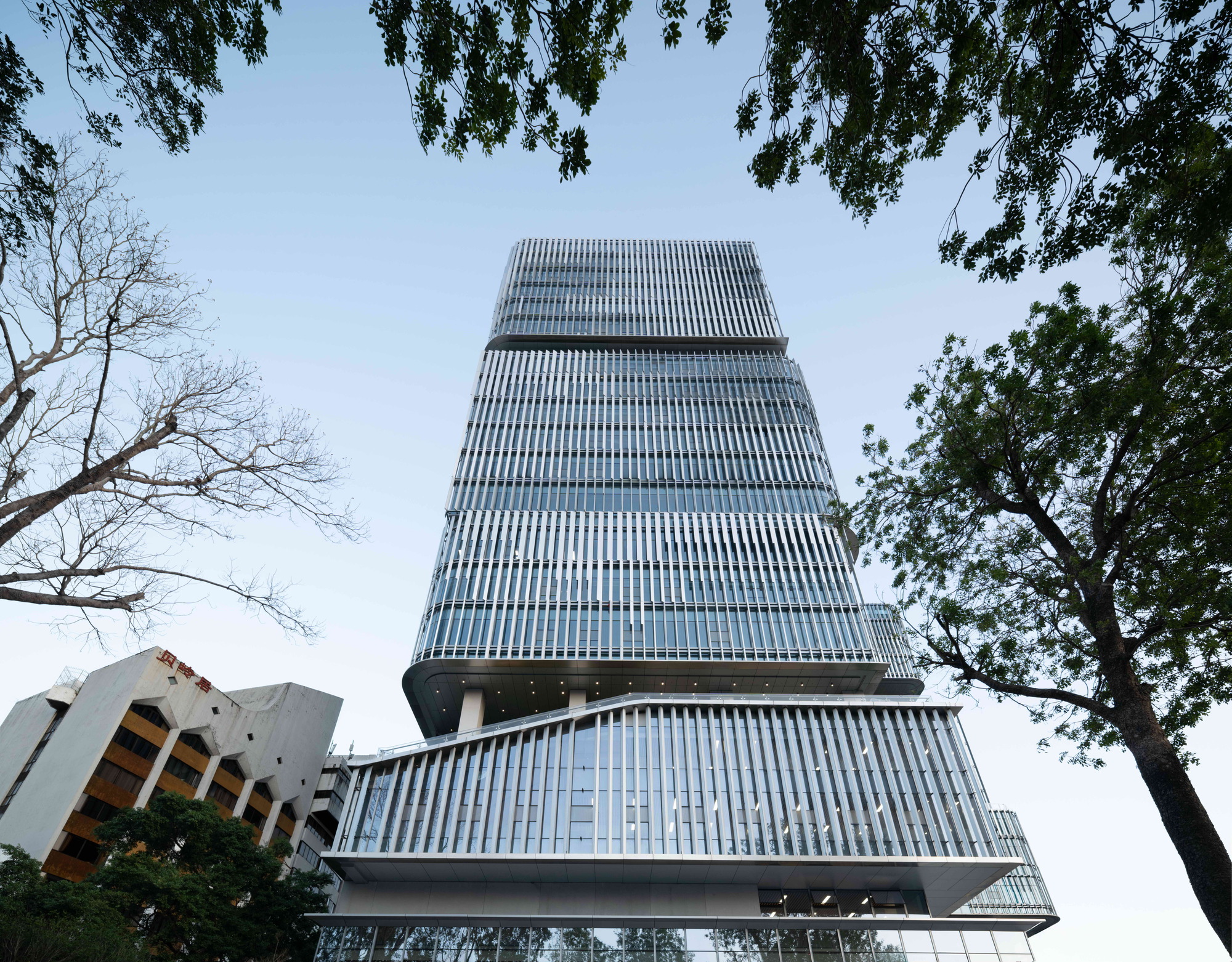 Low-angle view of the stacked tower with vertical fins, framed by tree branches against a pale dusk sky