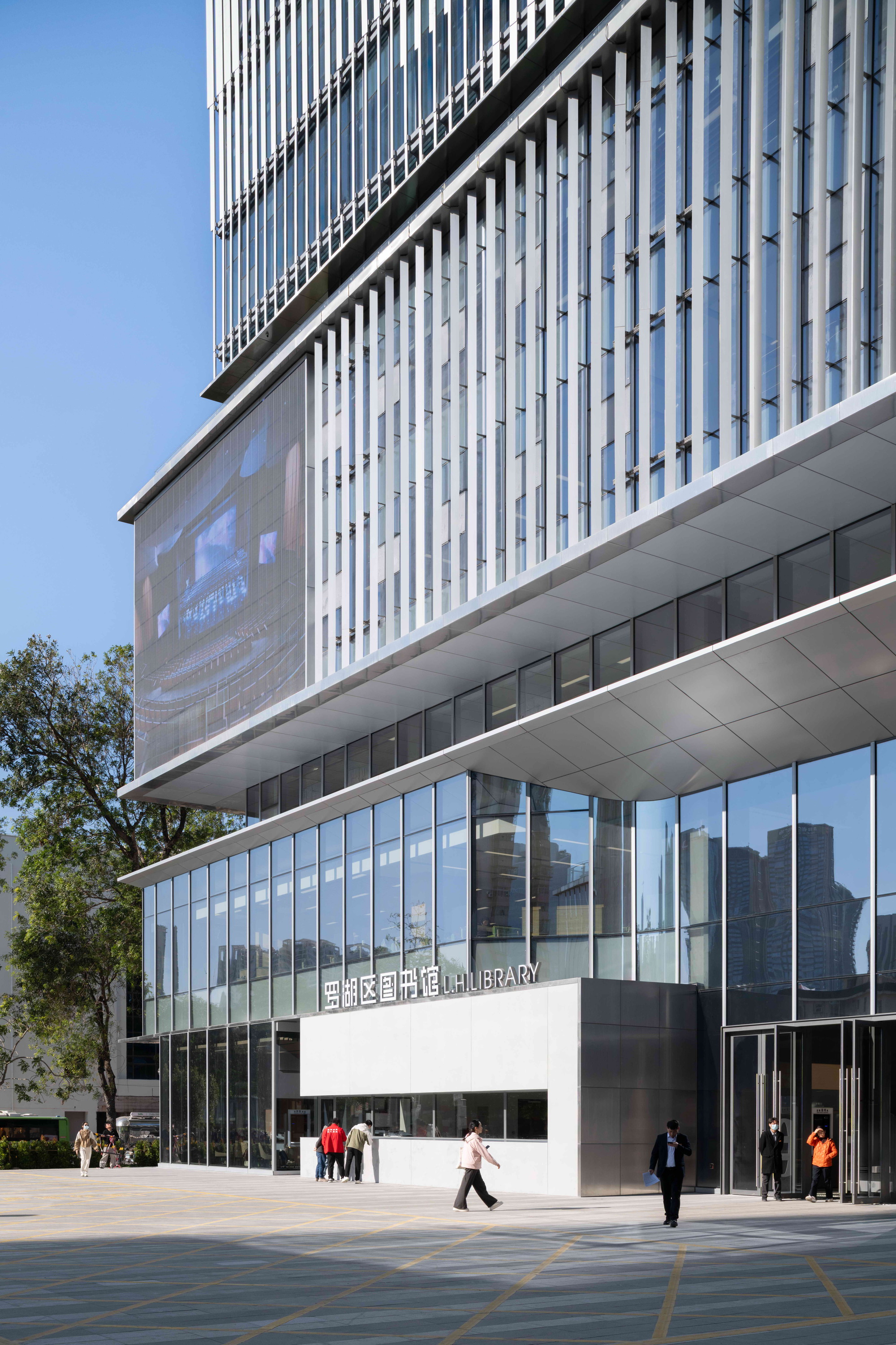 Library entrance below a projecting fin-clad volume and a large media screen, with people crossing the sunlit plaza