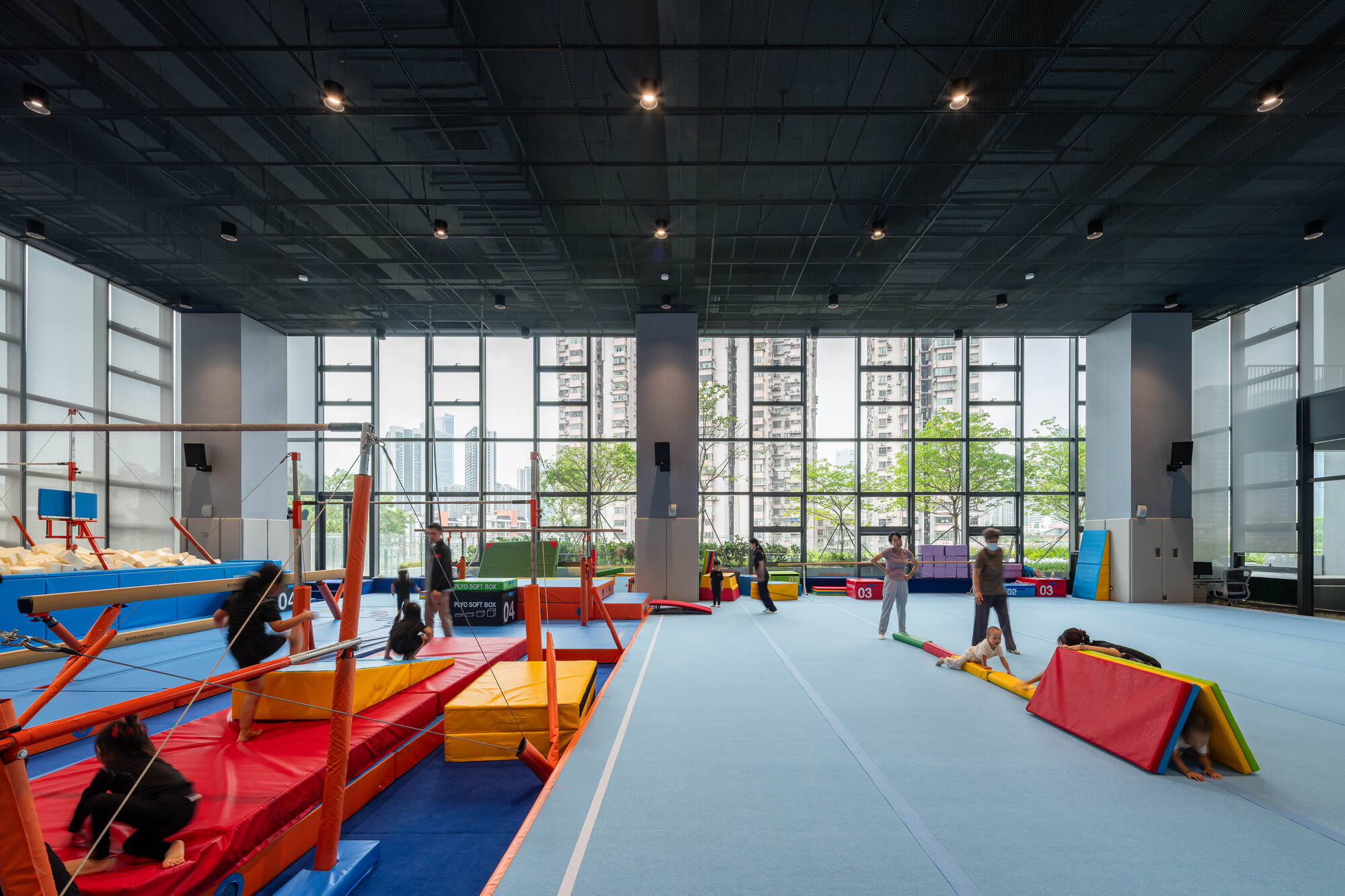 Indoor gymnastics hall with colored mats and bars, children playing beneath a black ceiling and tall windows facing apartment blocks