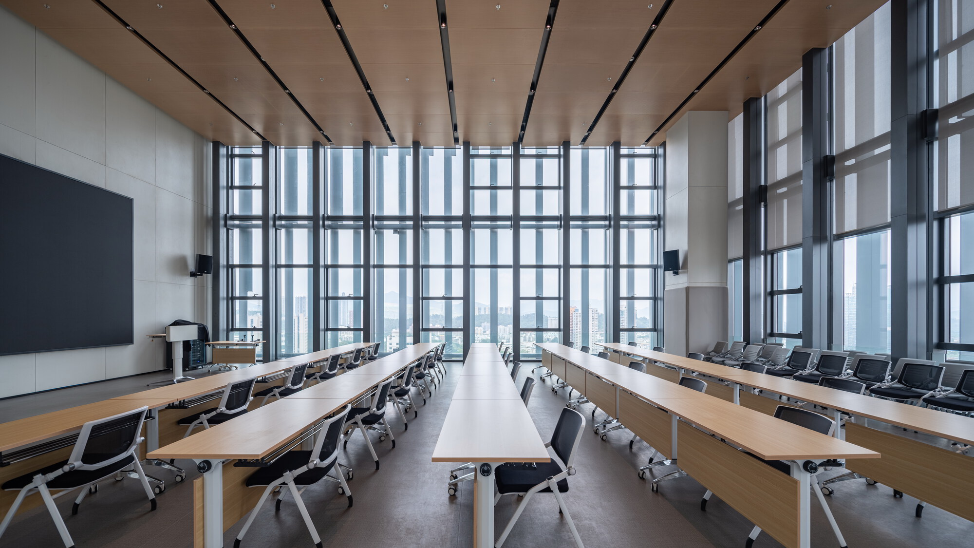 Training room with long timber desks and mesh chairs facing a full-height glazed wall with hazy city views