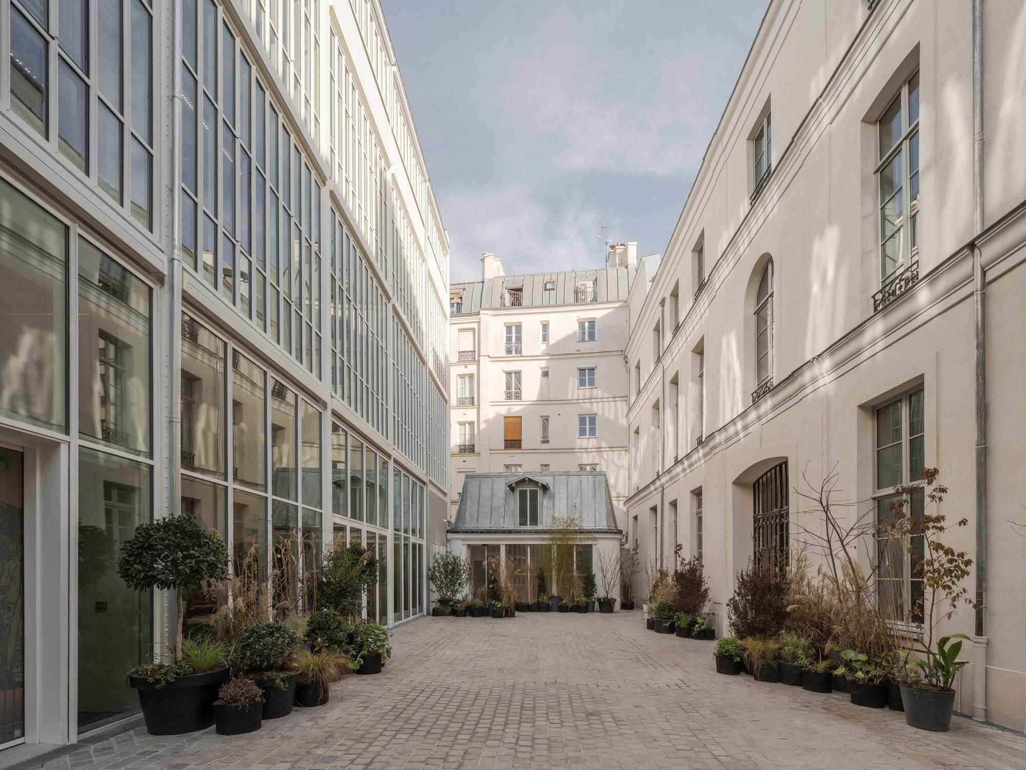 Cobbled courtyard between a glazed steel-framed facade and a plaster wing, with potted shrubs and a zinc-roofed pavilion at the far end
