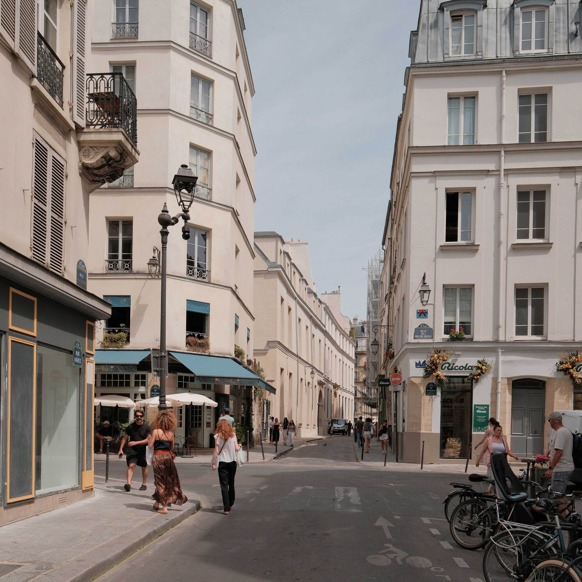 Street corner of cream stone apartment buildings with wrought iron balconies, a lamp post, a cafe awning, pedestrians and parked bicycles in summer sun