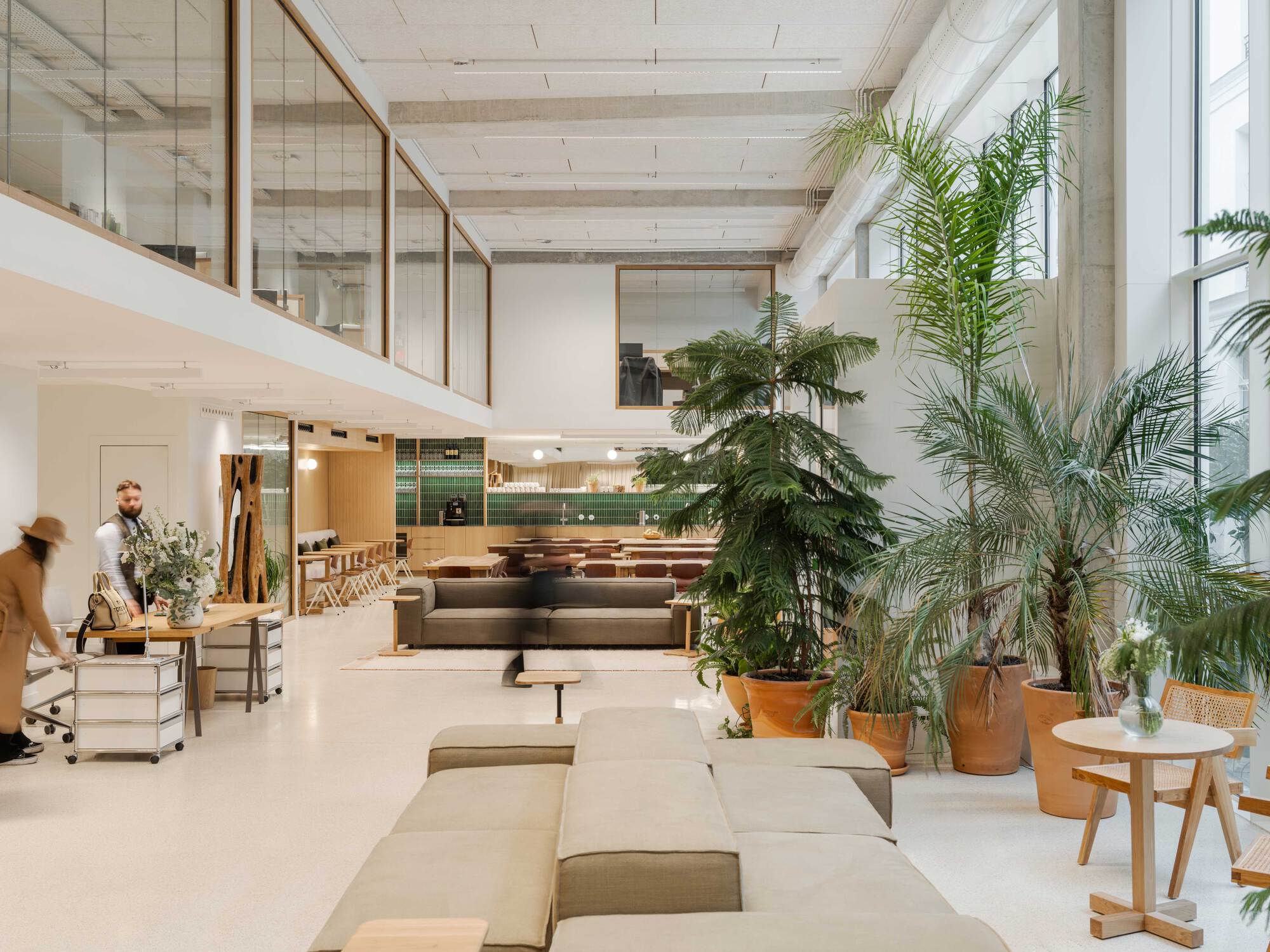 Double-height lounge with low modular sofas, potted palms in terracotta and a mezzanine behind timber-framed glazing, with two people at a table