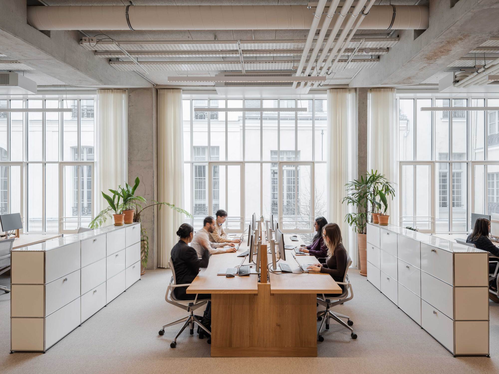 Open office with a shared oak desk, white storage units, concrete columns and exposed ducts, with staff working in front of tall curtained windows
