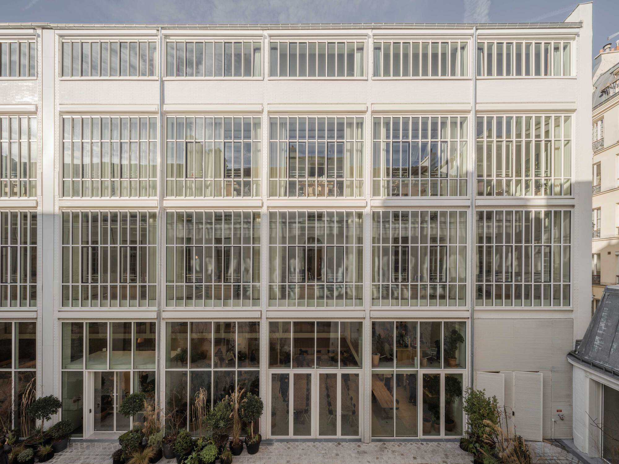 Frontal view of the white gridded glass courtyard facade rising four storeys above planters and a paved ground in clear light