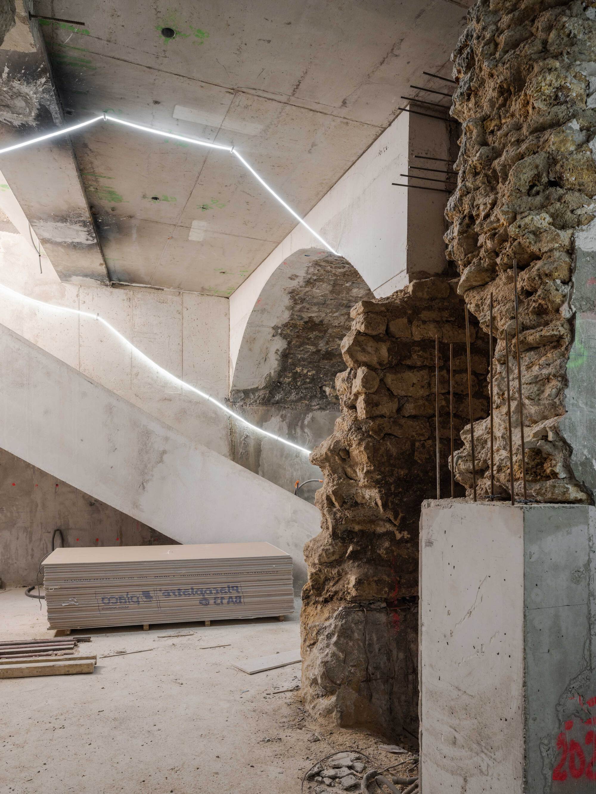 Construction-stage interior showing a rough rubble stone wall with rebar beside a concrete stair, outlined by a thin line of white light