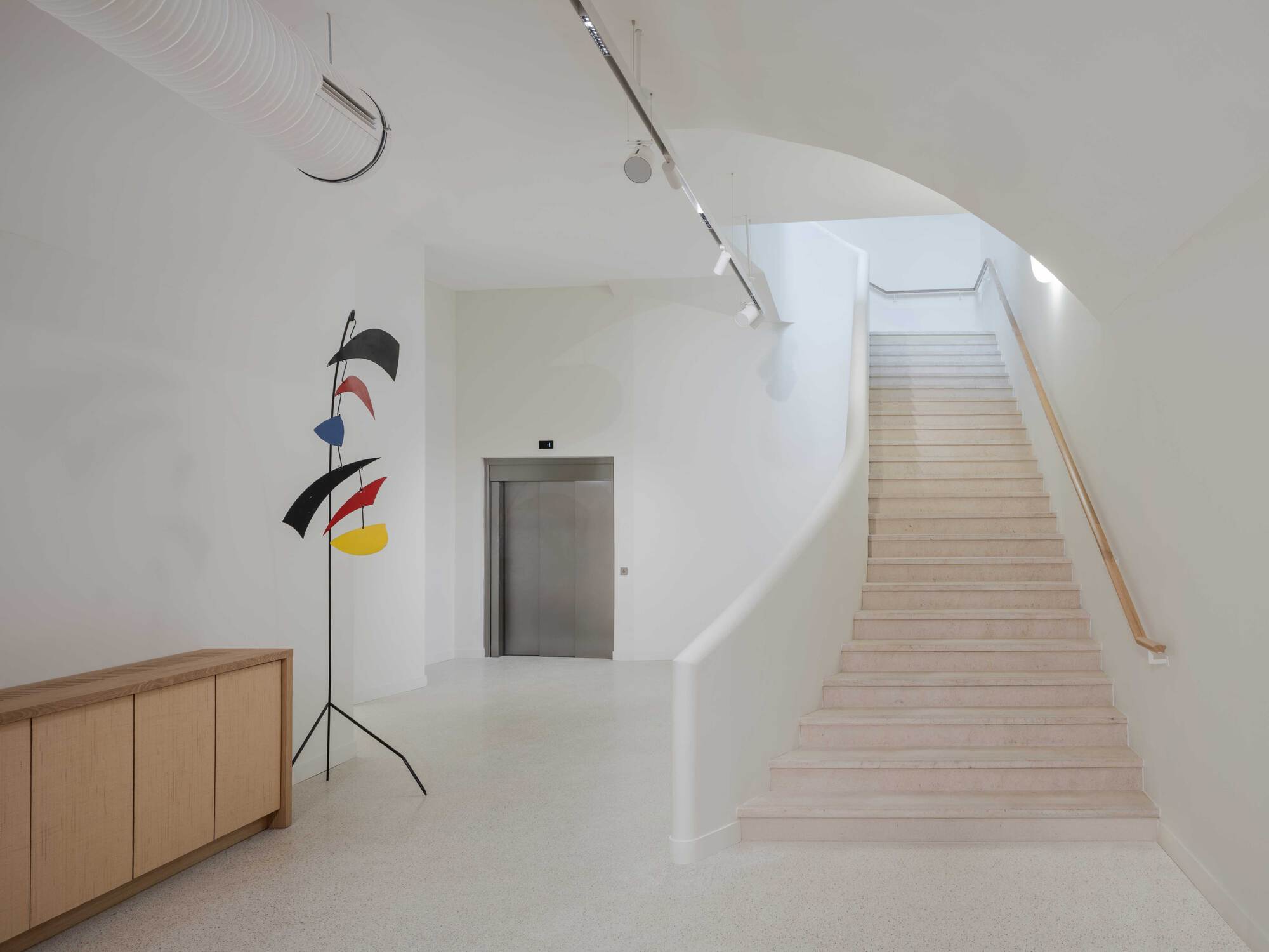 Finished stair hall with pale terrazzo steps, a curved white wall, a timber handrail and a hanging mobile sculpture beside an oak cabinet