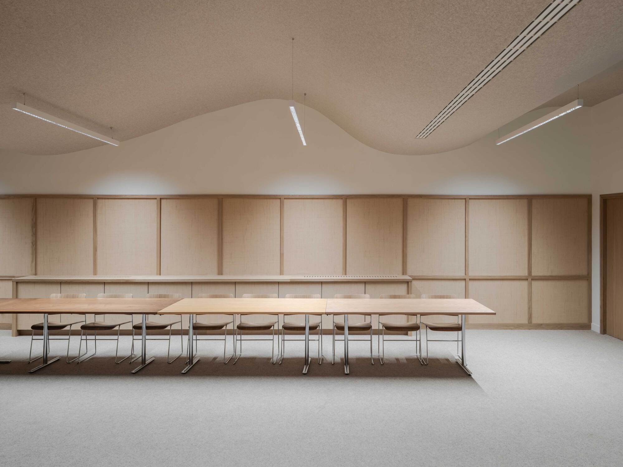 Long timber meeting tables and chrome-legged chairs before a paneled wall beneath a wavy ceiling edge and linear pendant lights