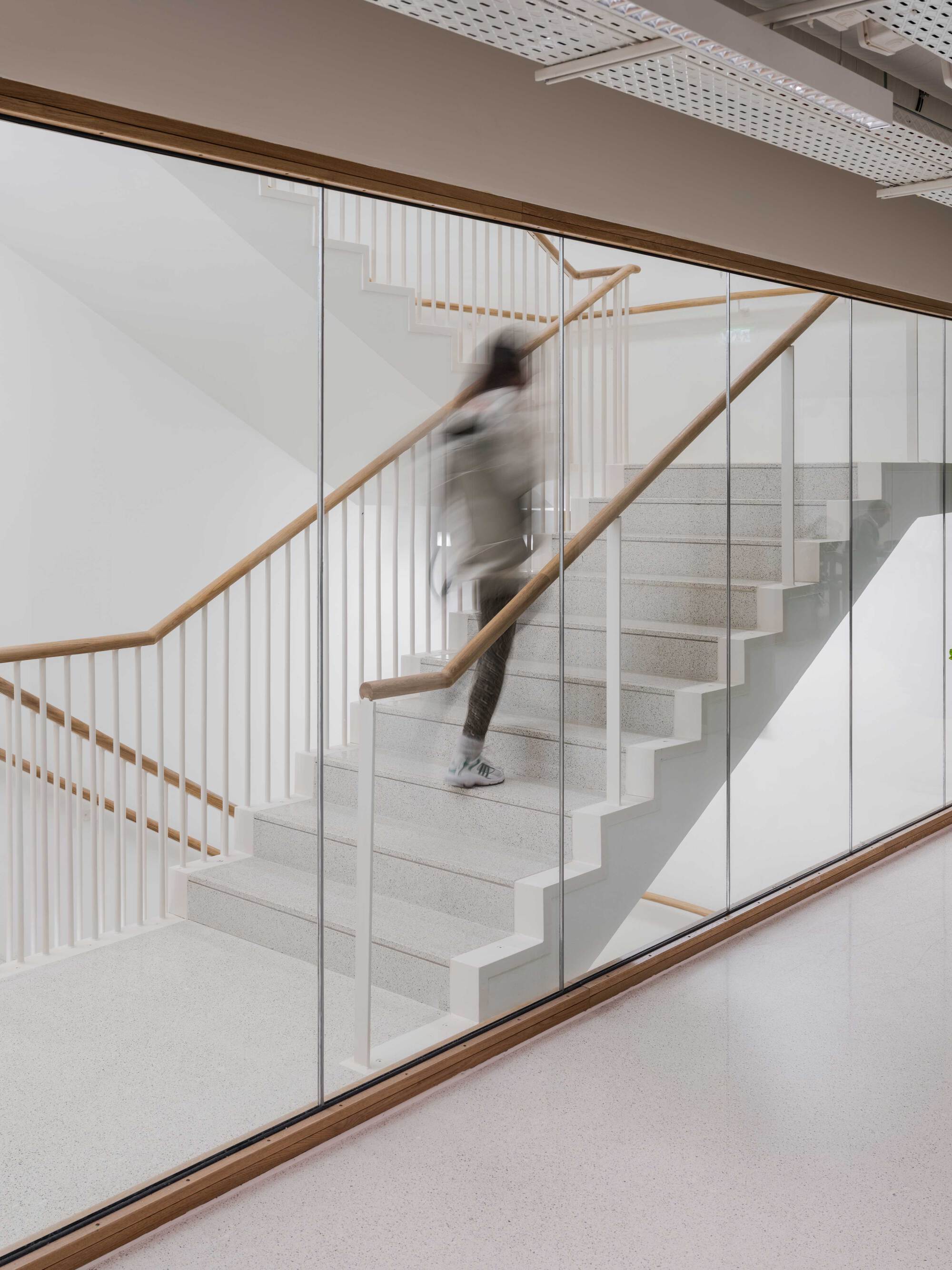 Blurred person climbing a white terrazzo stair with timber handrail, seen through a full-height glass wall