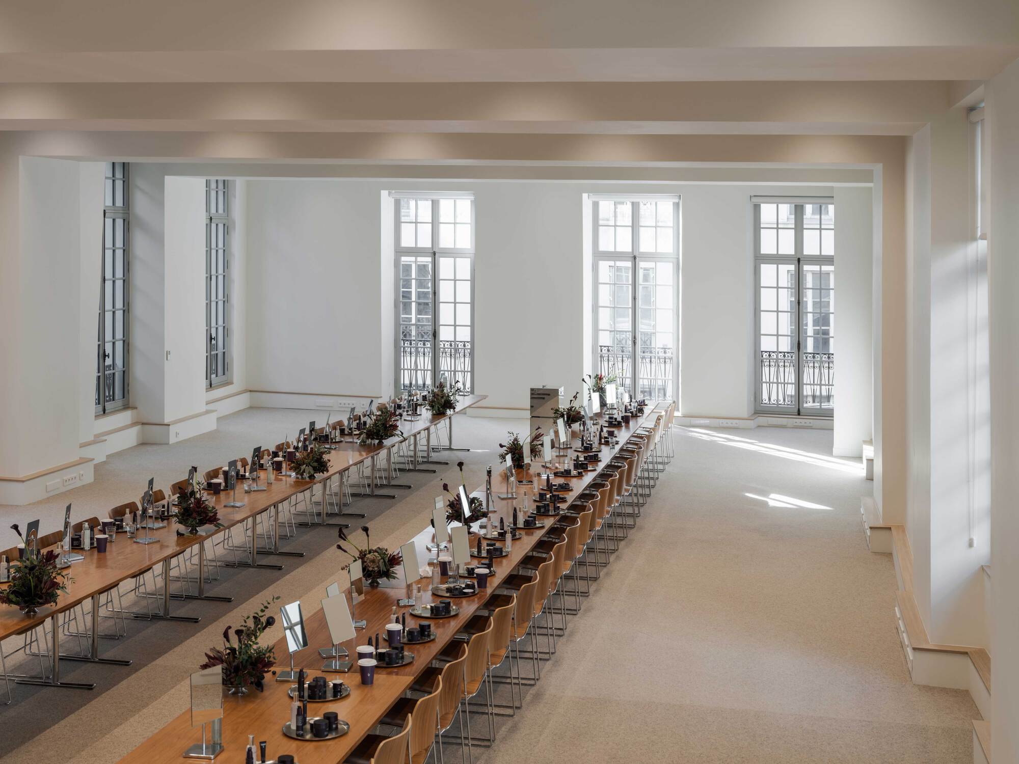 Long refectory tables with timber chairs and floral centerpieces beneath tall French windows, sunlight falling across the pale floor