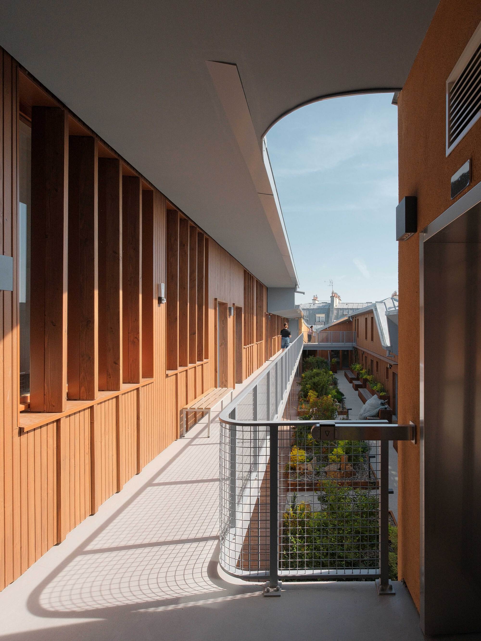Upper walkway beside vertical timber fins and a mesh balustrade overlooking a planted courtyard under a clear blue sky