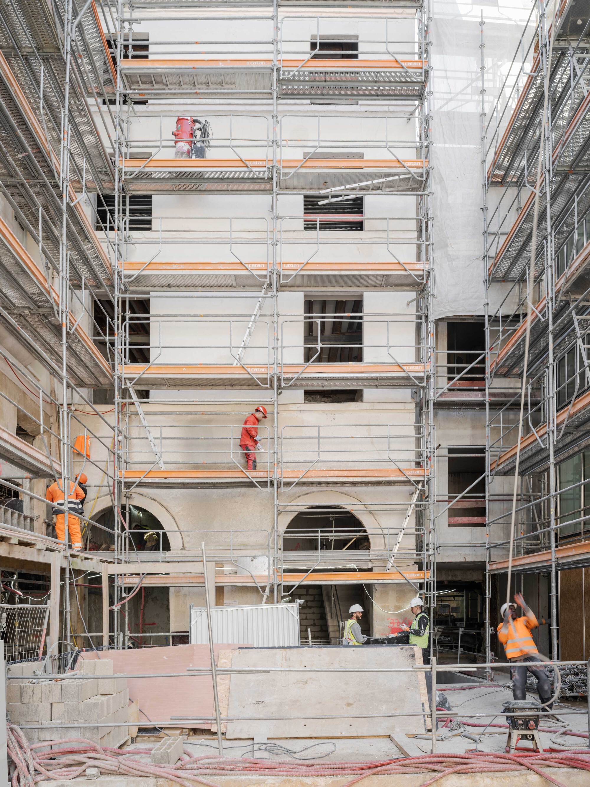 Scaffolded interior courtyard facade with workers in orange and an arched opening at its base during construction