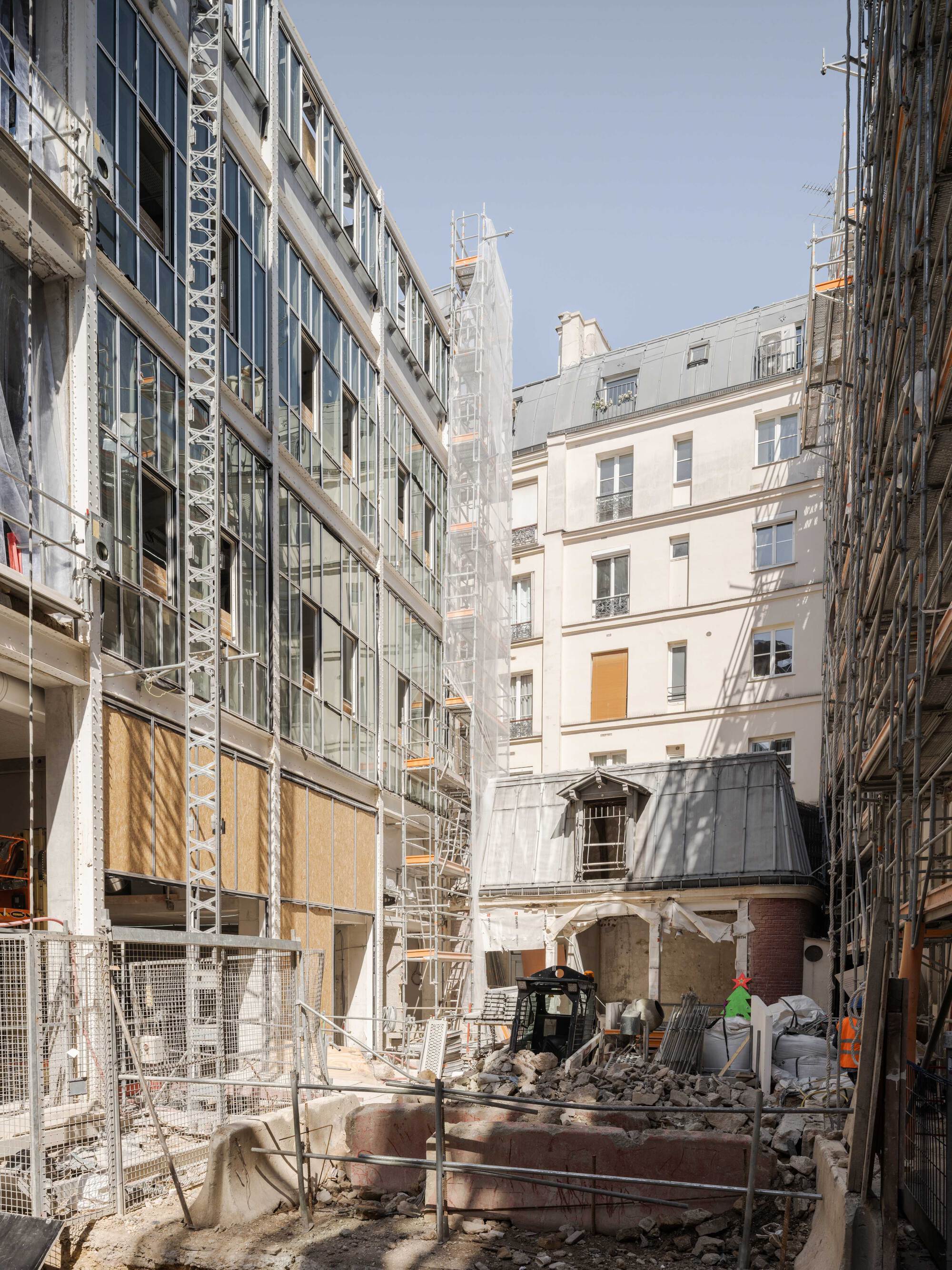 Glazed steel-frame facade beside a mansard-roofed masonry building, with rubble and scaffolding in the foreground on a sunny day