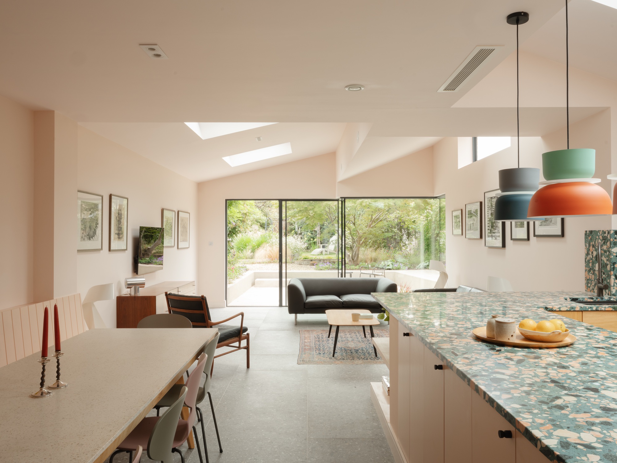 Open-plan kitchen and living room with terrazzo island, pendant lamps and sliding glass doors opening onto a leafy garden in daylight
