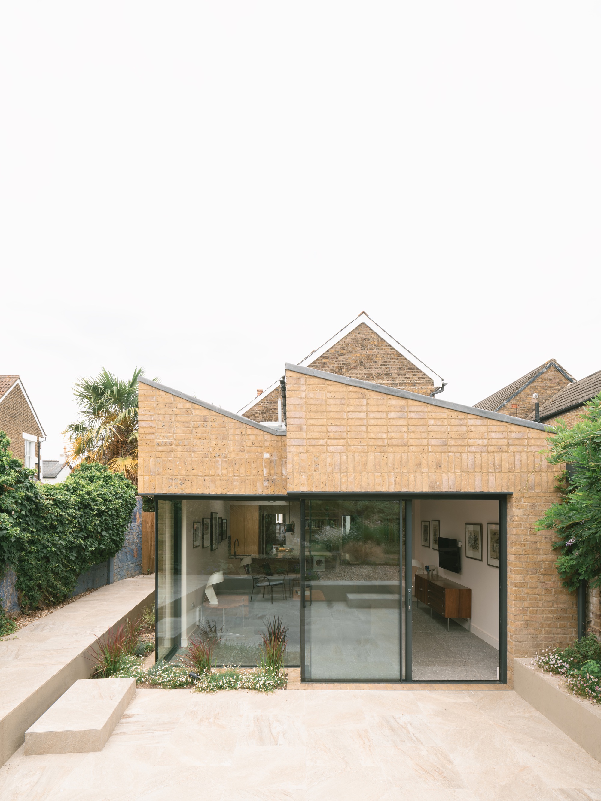 Rear brick extension with two sloping parapets and sliding glazed doors facing a pale stone terrace under an overcast sky
