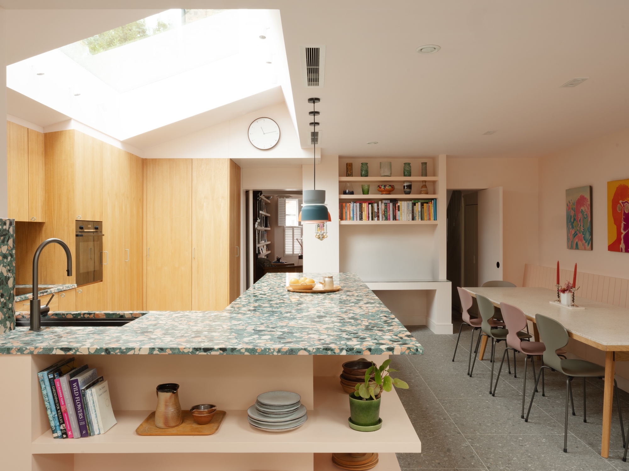Terrazzo-topped kitchen peninsula with open shelving, oak cabinets and a long dining table beneath a large rooflight