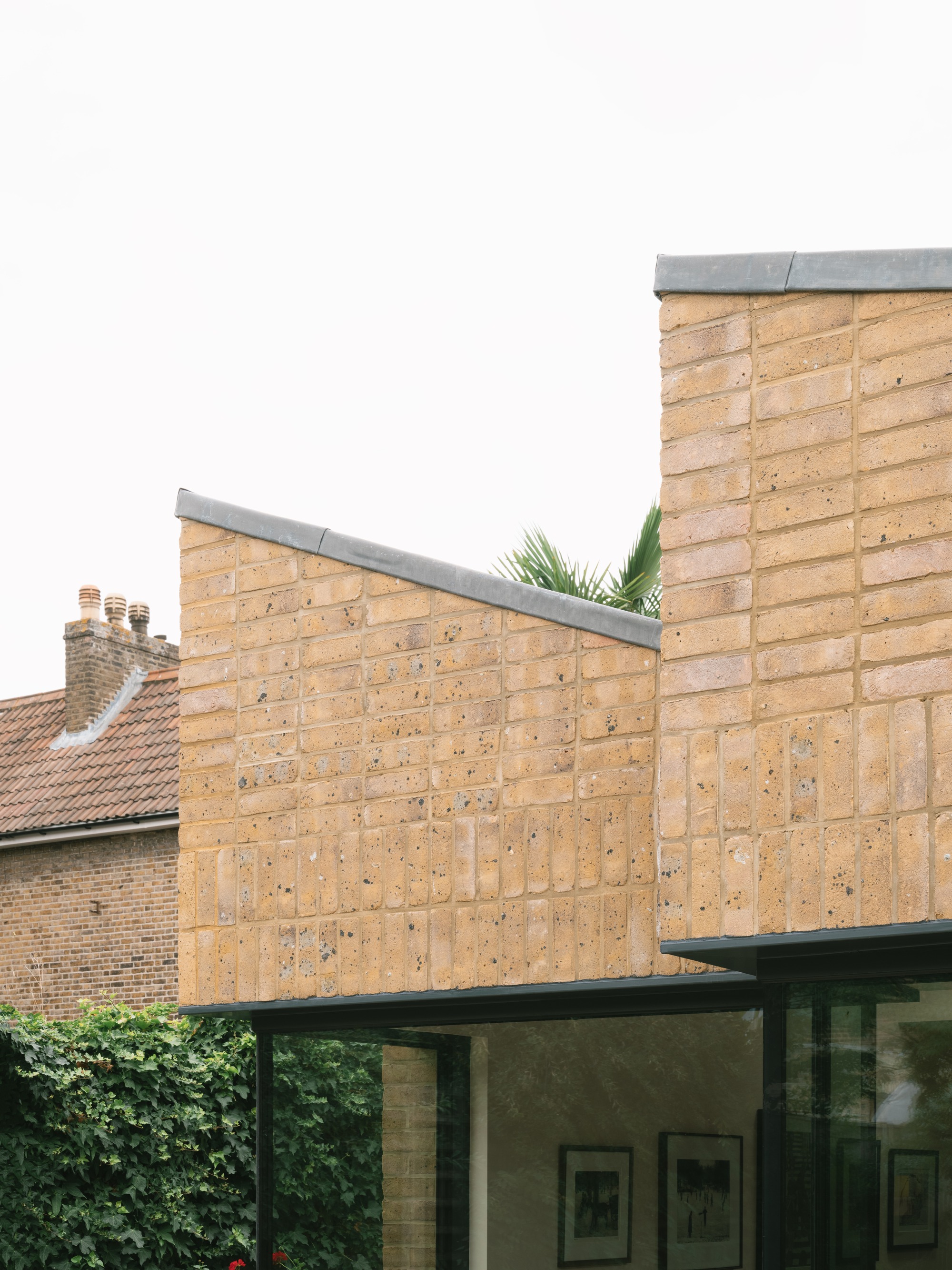 Close-up of yellow brick parapet walls with slanted zinc capping, set against a neighbouring tiled roof and ivy