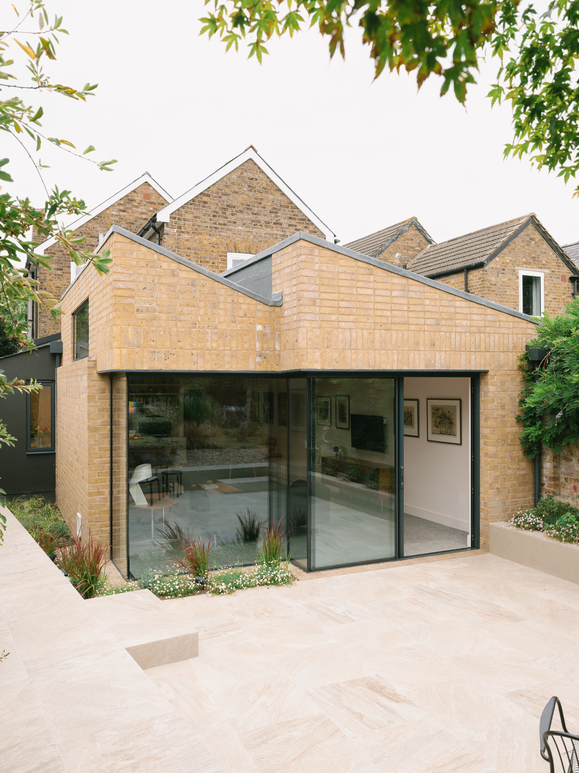 Garden view of the brick extension with corner sliding glazing, raised planter and stone paving under overhanging tree leaves