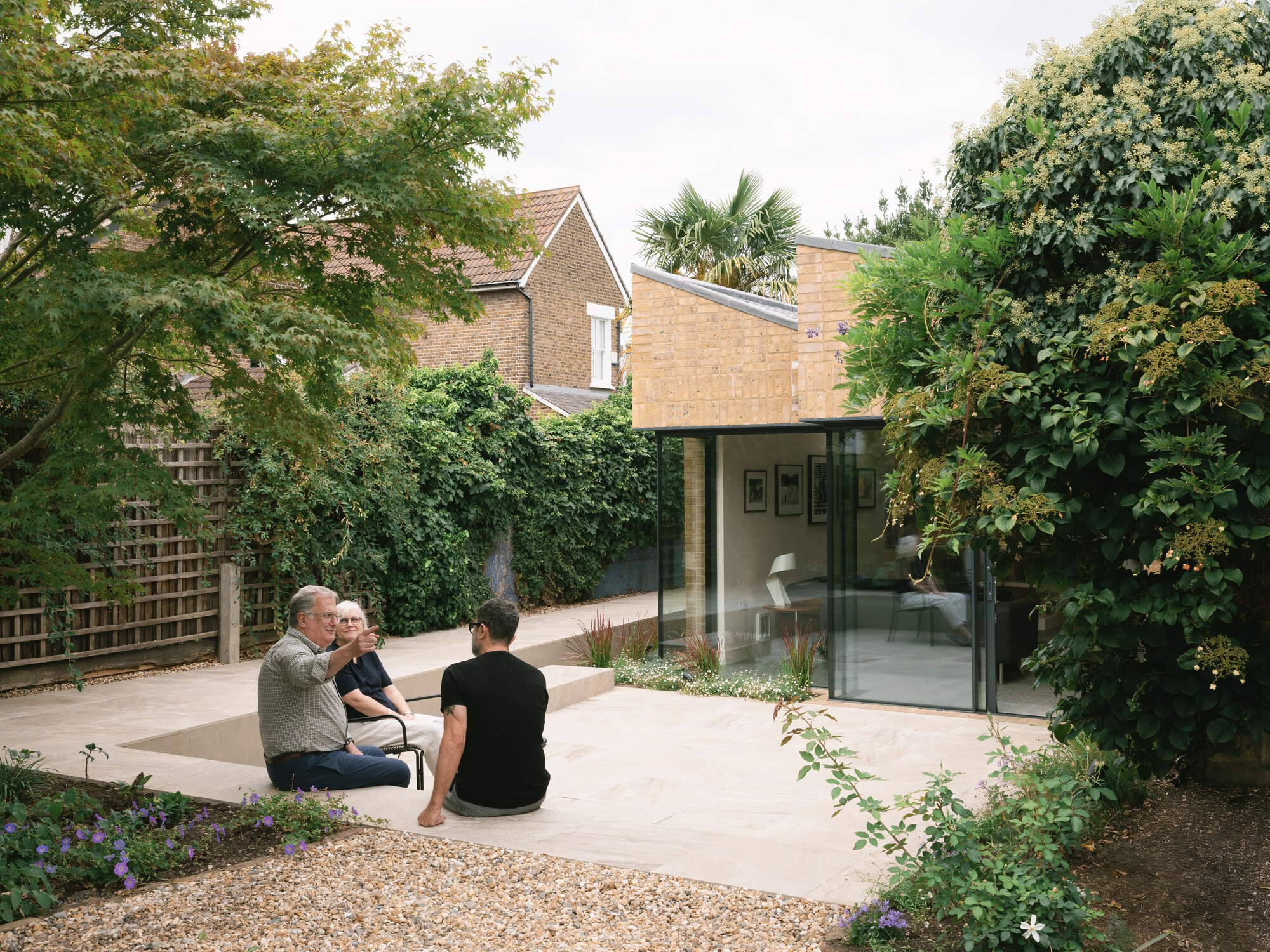 Three people seated on a pale stone terrace facing a glazed brick garden extension, framed by climbing plants and a maple