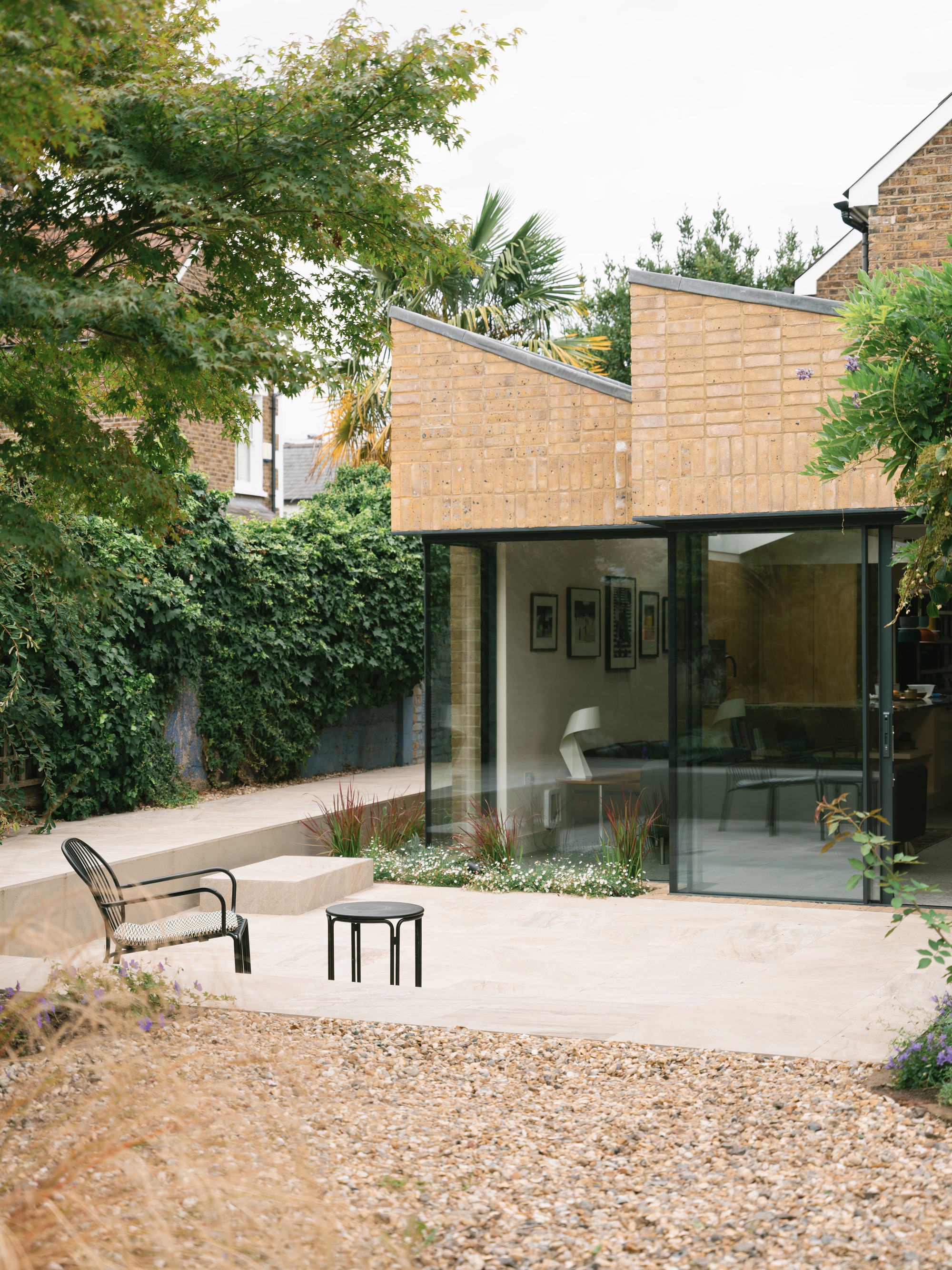 Yellow brick extension with stepped sloping parapets and corner glazing, seen over a gravel bed and stone steps with a black chair