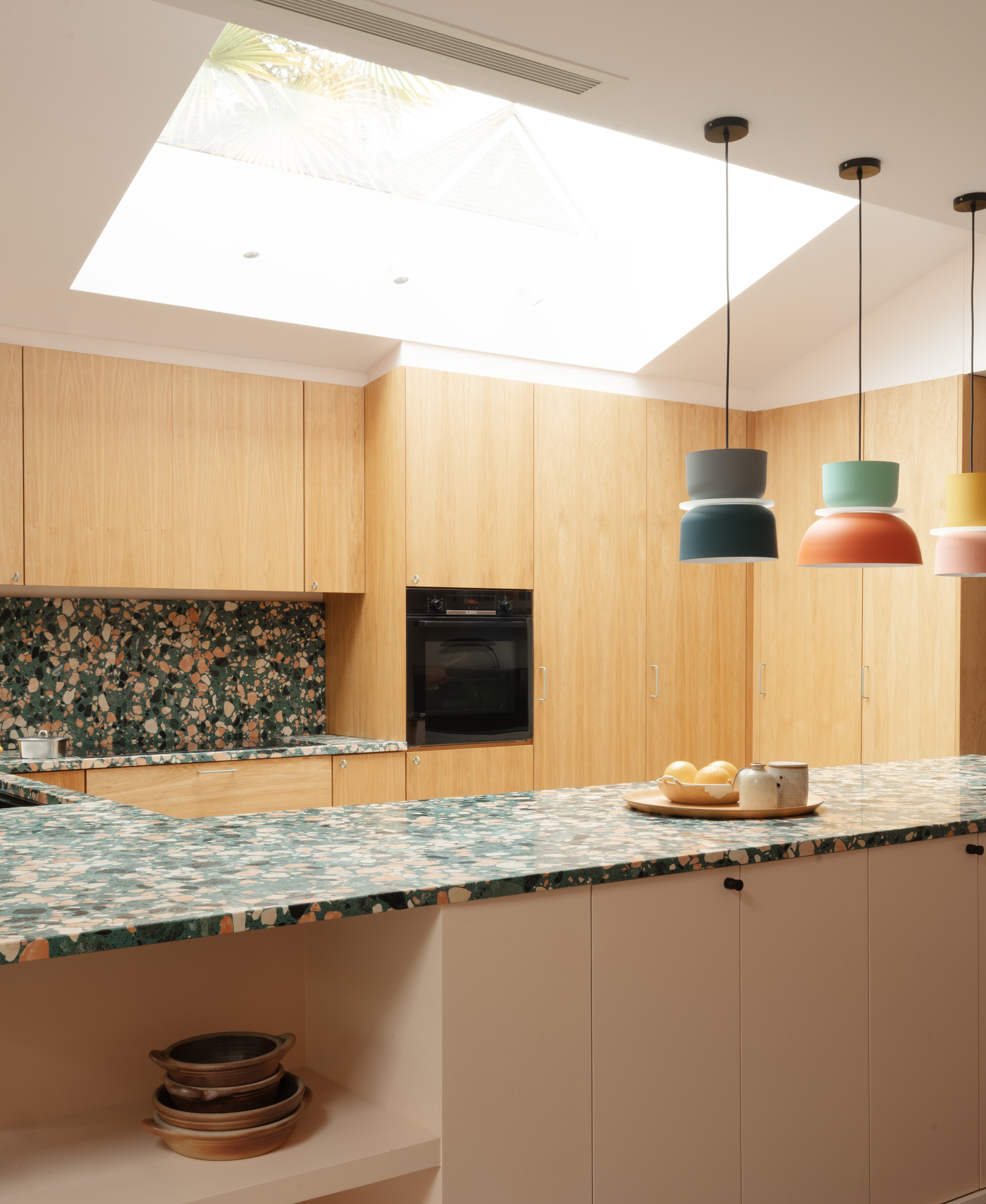 Kitchen island with green terrazzo worktop beneath a large rooflight and three colourful pendant lamps over oak cabinetry