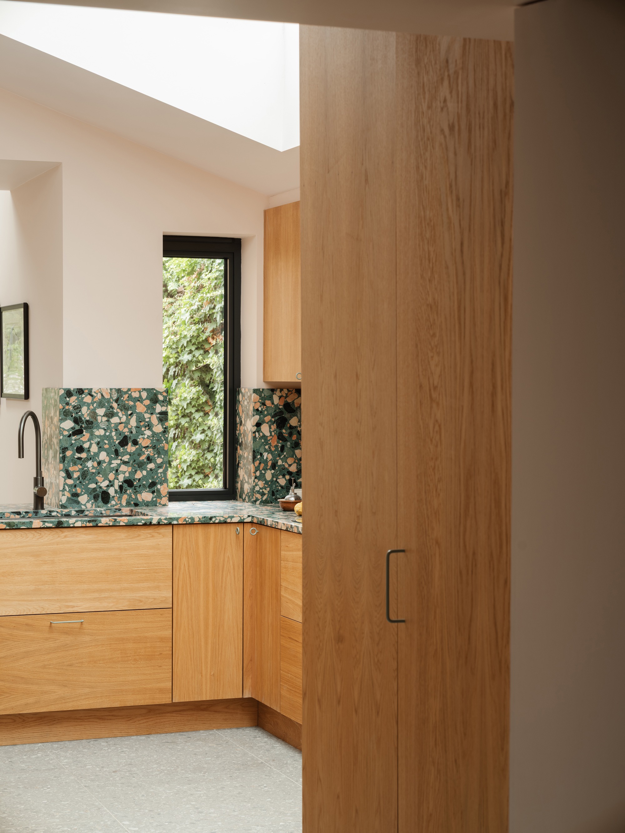 Oak kitchen units with terrazzo splashback and a narrow window onto ivy, glimpsed past a tall timber door in soft daylight