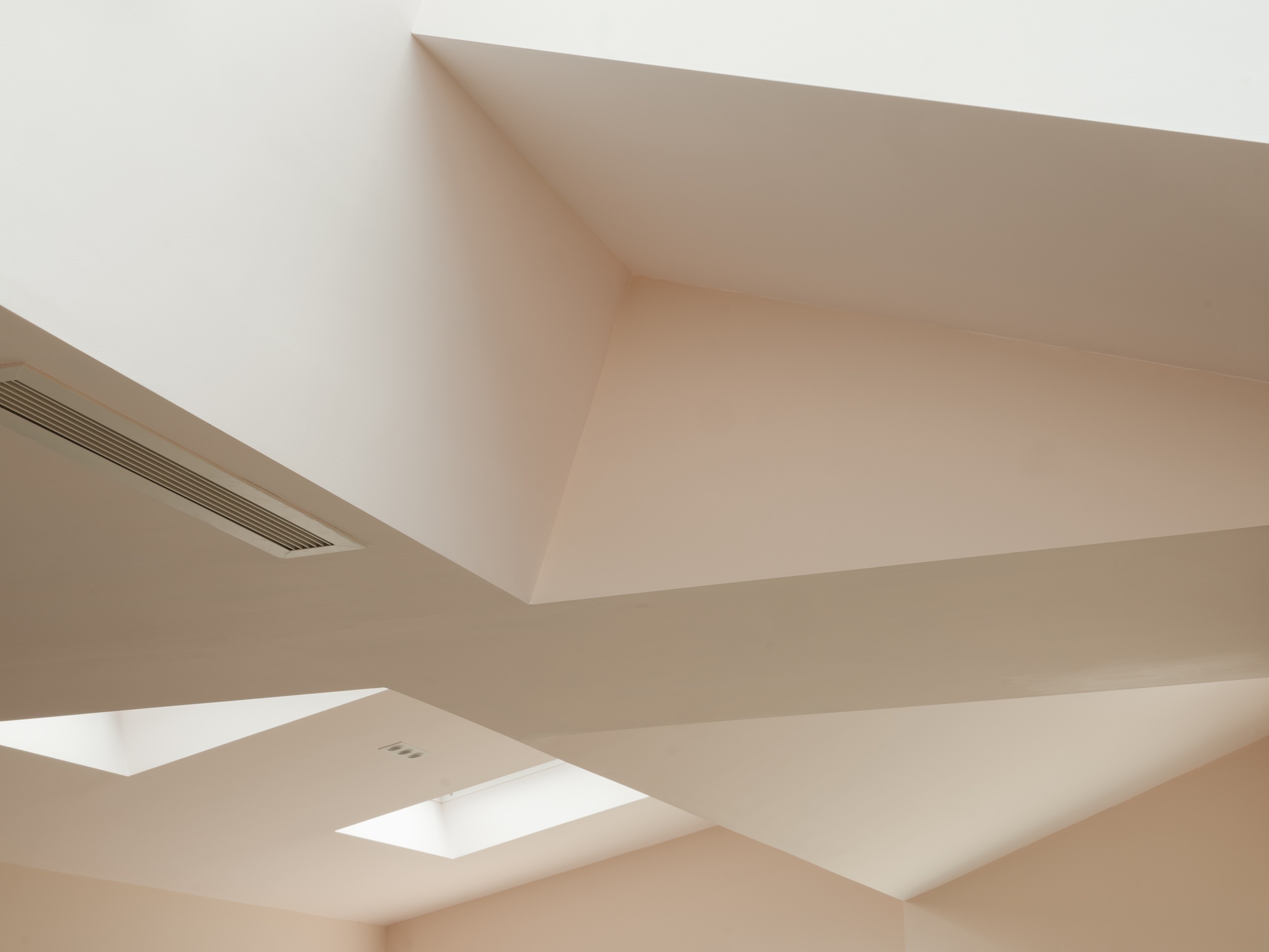 Upward view of the faceted white plaster ceiling with angled planes, a linear air vent and two small skylights