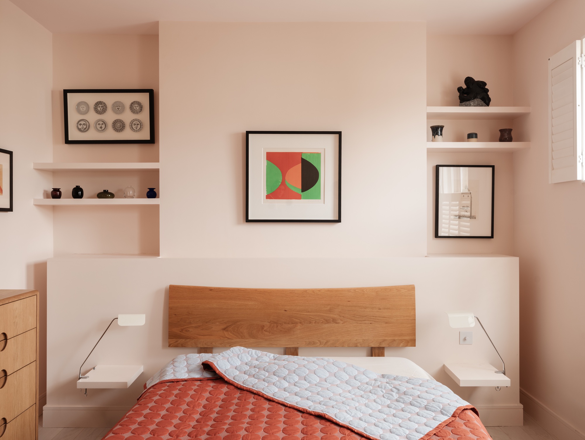 Bedroom with an oak headboard against a pale pink niche wall, framed prints, small shelves and an orange quilted bed cover in soft daylight