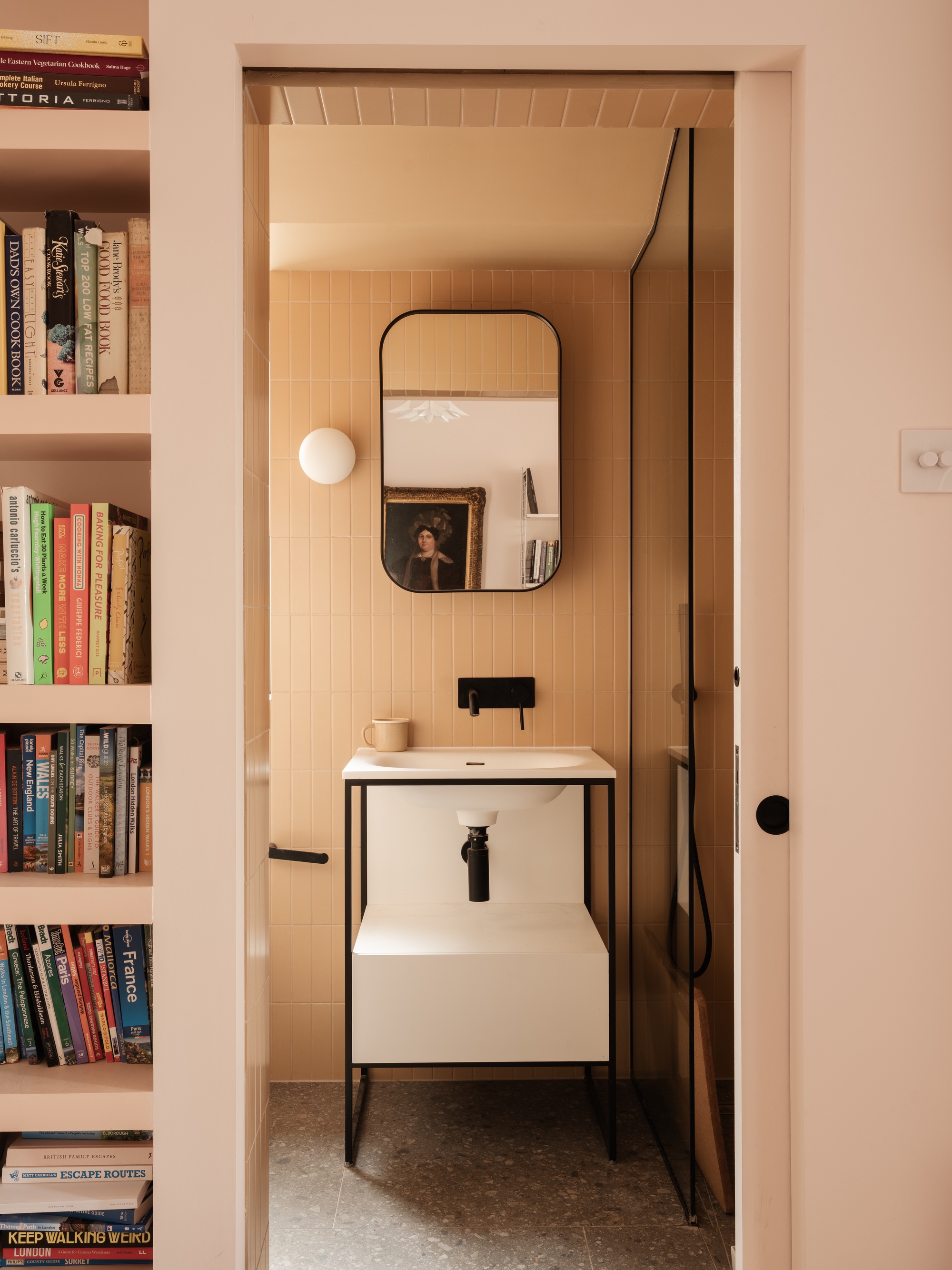 Pale pink bathroom glimpsed through a doorway beside a bookshelf, with vertical tiles, a black-framed mirror and a freestanding white basin
